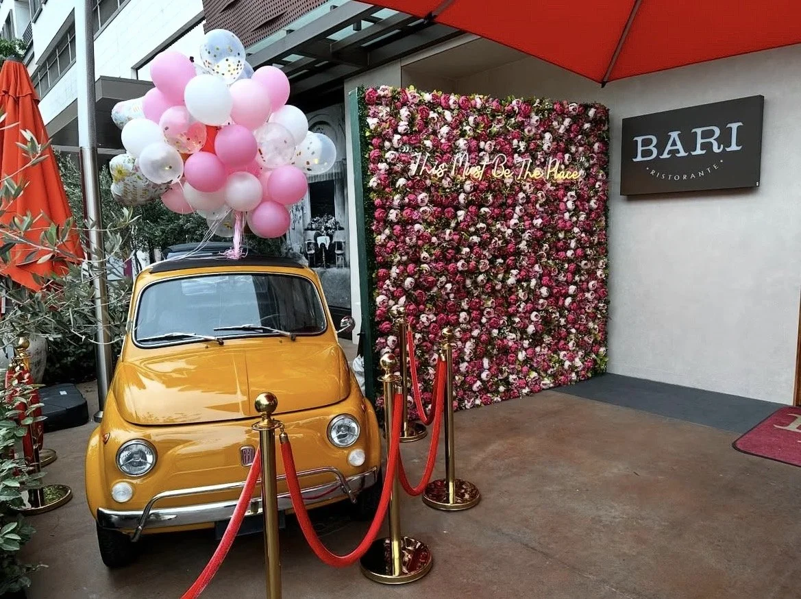 A yellow vintage Fiat car with pink, white, and gold balloons on top, roped off with red velvet ropes and gold stanchions, is parked next to a flower wall with pink and white flowers at a restaurant entrance.