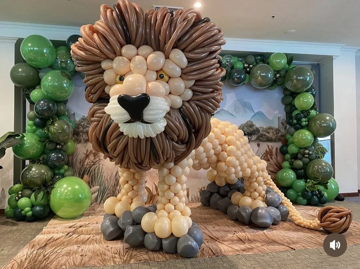Balloon sculpture of a lion with a detailed face, mane, and body, surrounded by green and black balloons, set against a nature-themed backdrop.