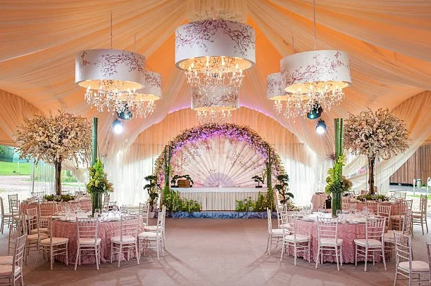 Elegant event tent decorated for a celebration with round tables, pink tablecloths, white chairs, large floral centerpieces, chandeliers, and a floral archette at the front.