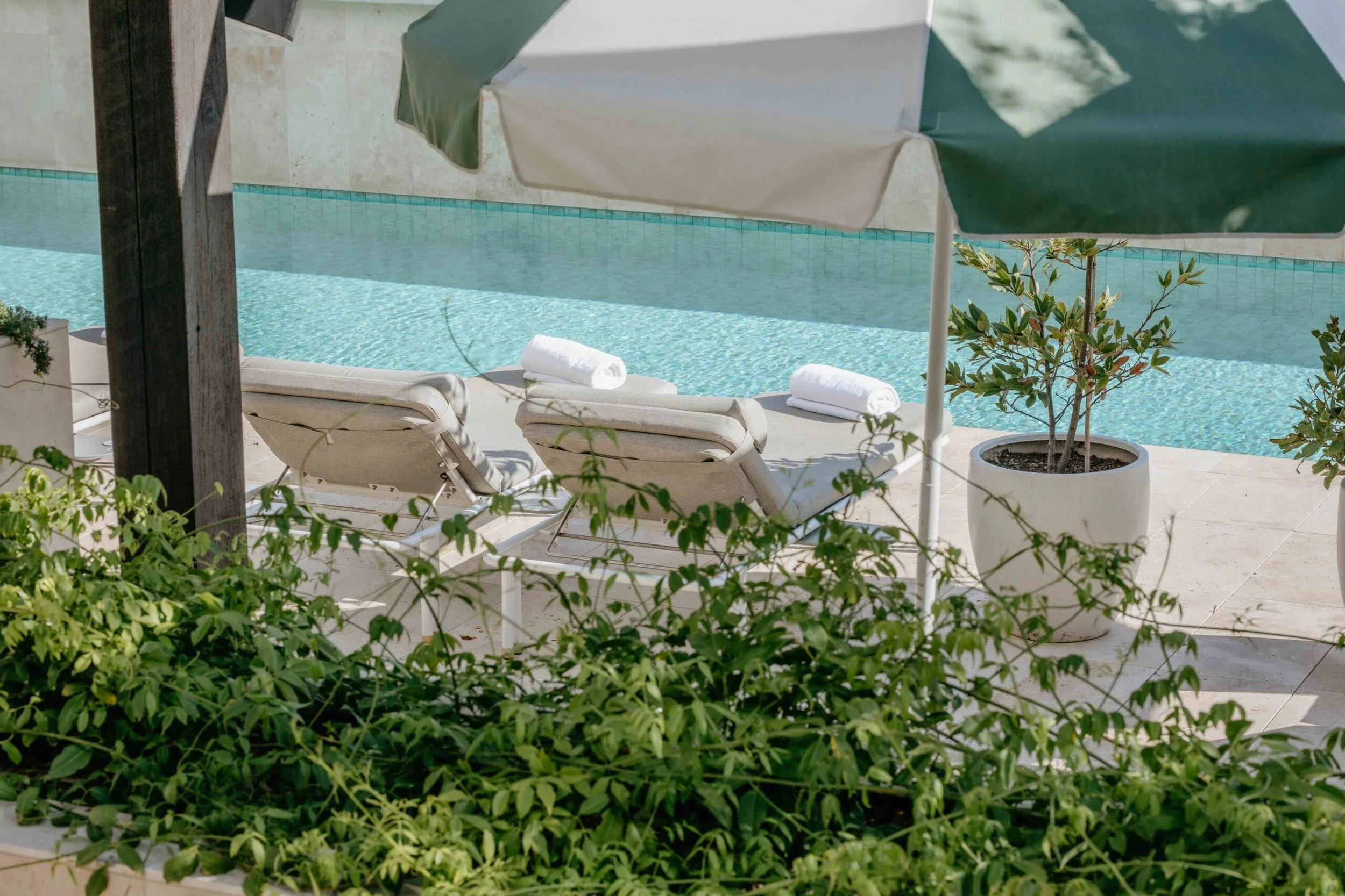 Poolside view with two lounge chairs, rolled white towels, a large green umbrella, potted plants, and a swimming pool with blue tiles.