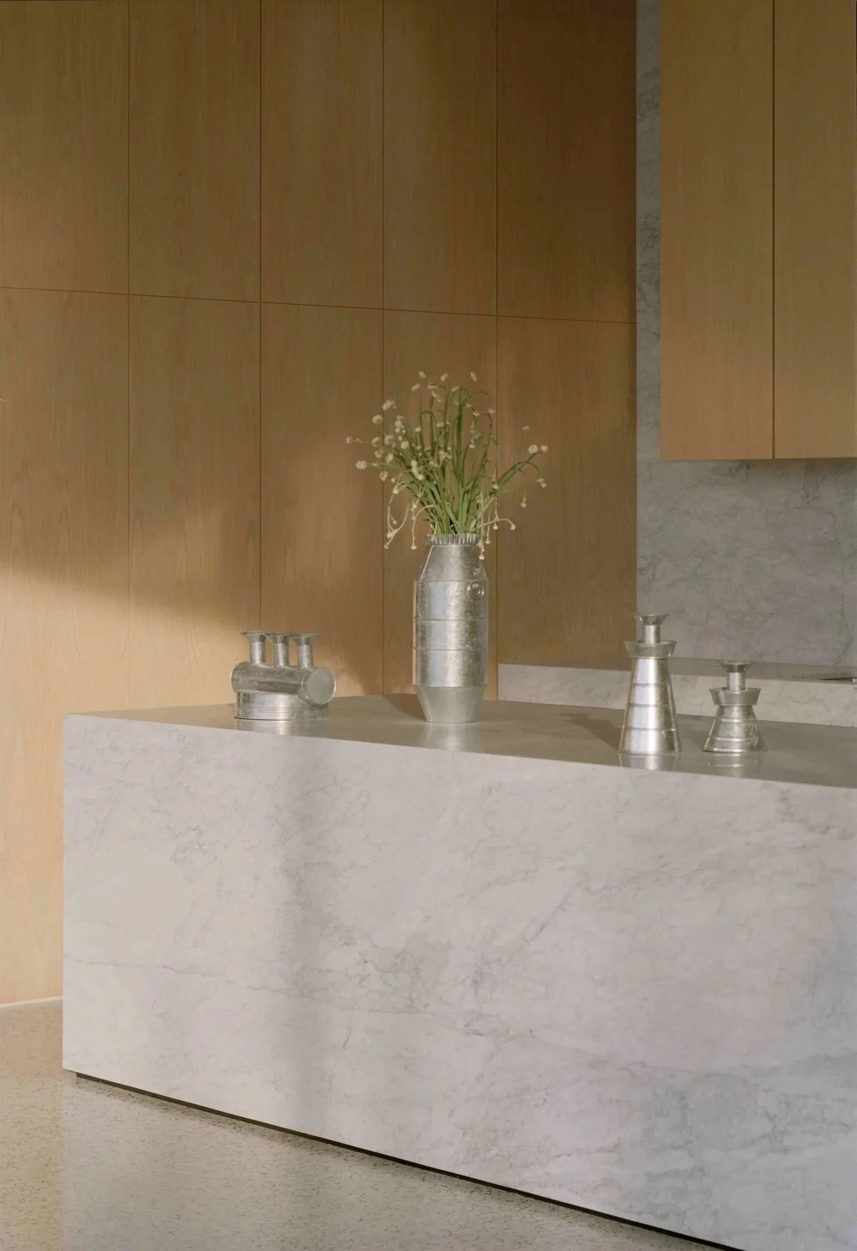 A minimalist interior with a white marble countertop, a metal vase with white flowers, and metal fixtures against a wooden wall and a marble backsplash.