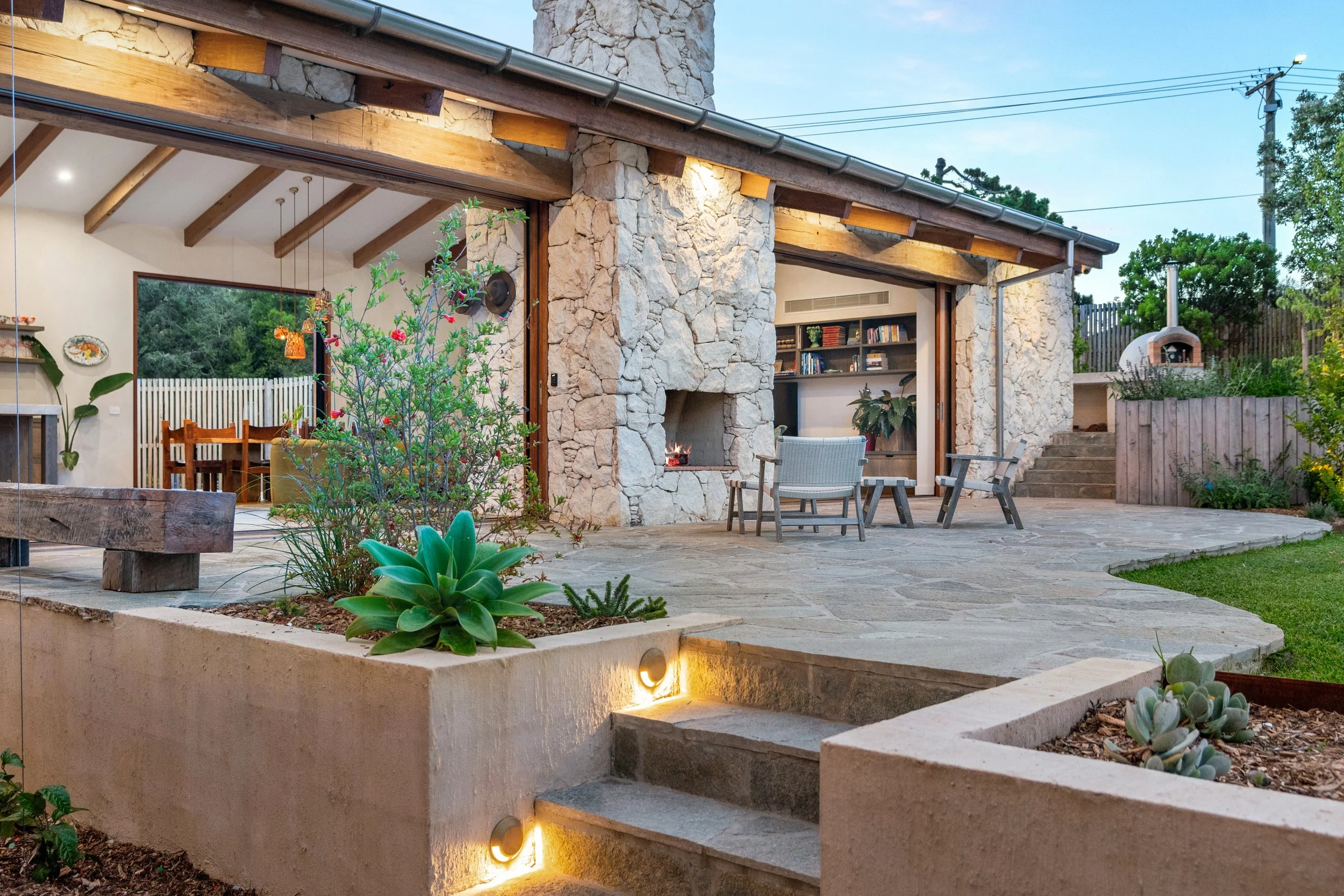 A backyard patio area with stone walls, wooden beams, and outdoor furniture including chairs and a fireplace. There are plants and greenery, and a pizza oven is visible in the background. The patio is illuminated with soft lighting at dusk.