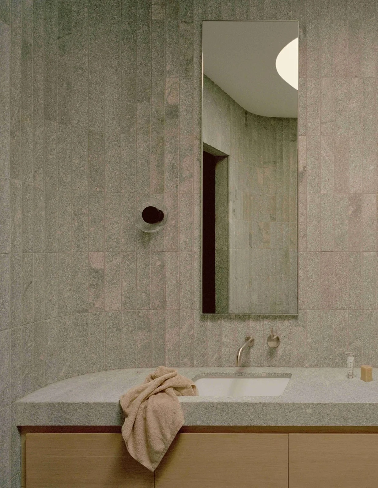 Modern bathroom with curved tiled wall, a rectangular mirror, a sink with a faucet, a soap bar, and a towel draped over the countertop. The mirror reflects the ceiling and a partial wall.
