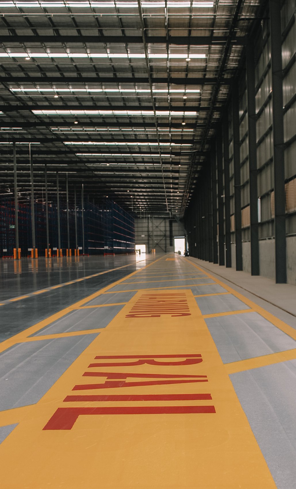 Empty warehouse with yellow and red painted markings on the floor, large open space, high ceiling with metal beams, and tall shelves along one side.