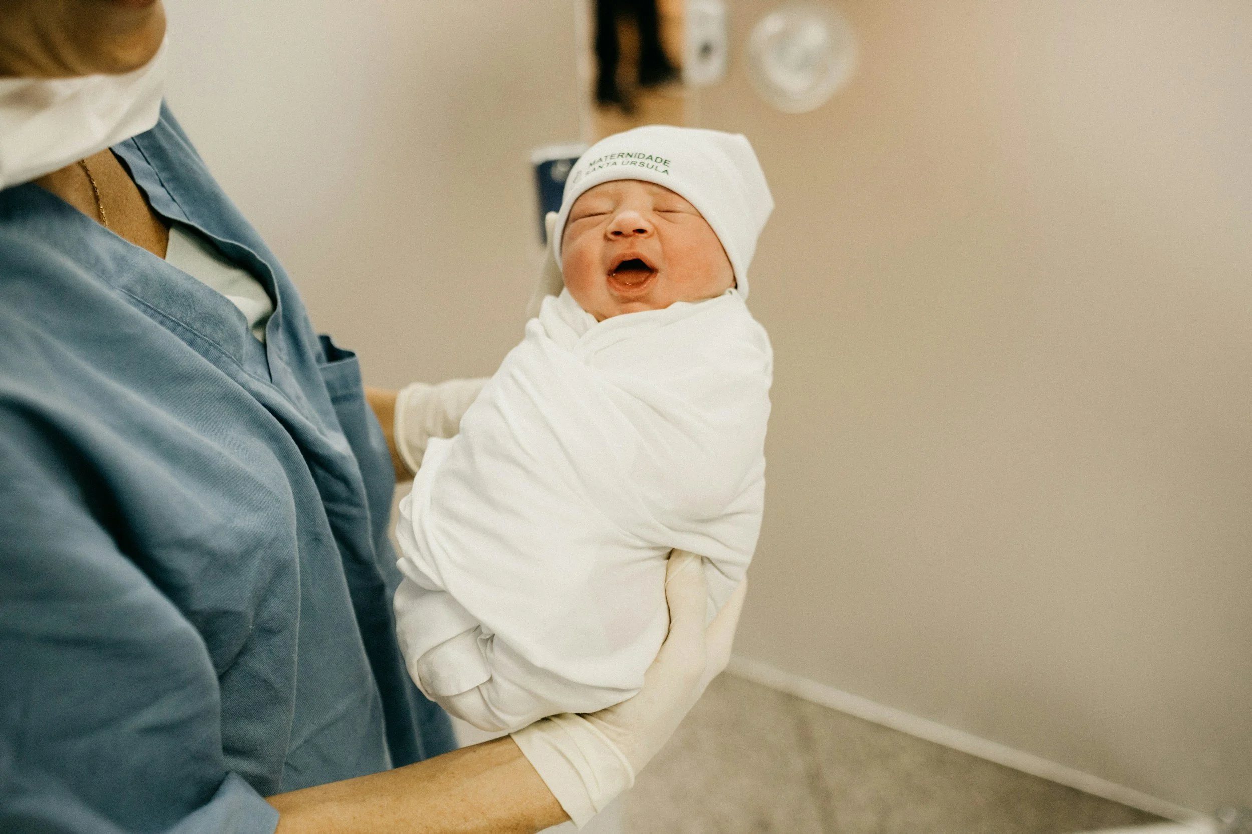 A newborn baby wrapped in a blanket is being held by a person in a hospital room.