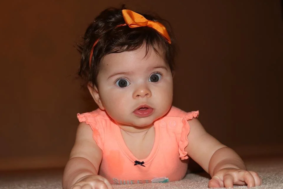 A surprised baby girl with curly dark hair secured with an orange bow, wearing a coral sleeveless top, lying on her stomach on a beige carpet with a brown background.