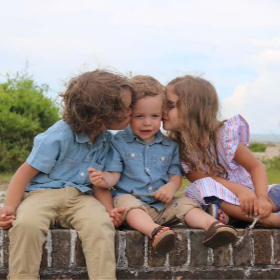 Two young children, a boy and a girl, kissing a younger child on the cheeks while sitting on a brick ledge outdoors, with green trees and a cloudy sky in the background.