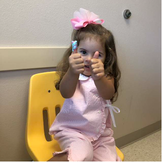 A young girl with long brown hair wearing a pink and white outfit and a large pink bow sits on a yellow chair giving a thumbs-up.