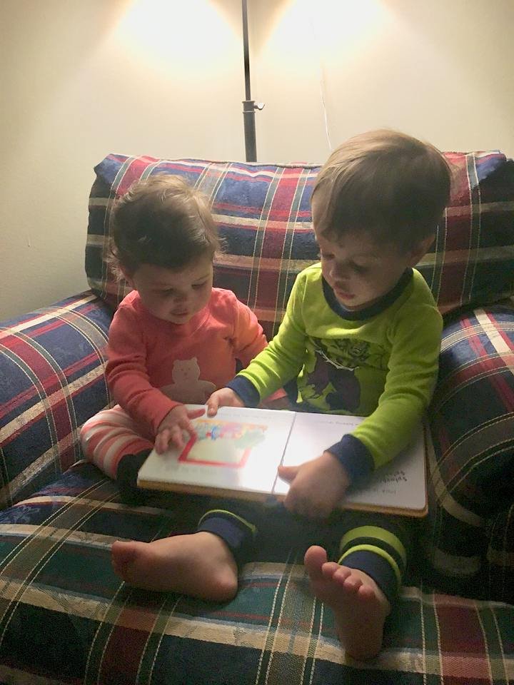 Two young children sitting on a plaid couch, looking at a picture book together. The boy is wearing pajamas with Batman and the girl is wearing striped pajamas. They are illuminated by a tall floor lamp behind them.
