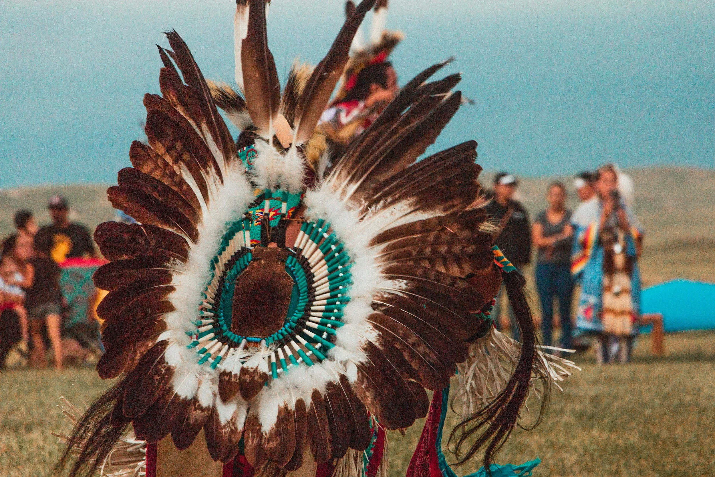 Close-up of a Native American headdress with brown and white feathers, adorned with turquoise beads, during a cultural gathering outdoors with blurred group of people in the background.