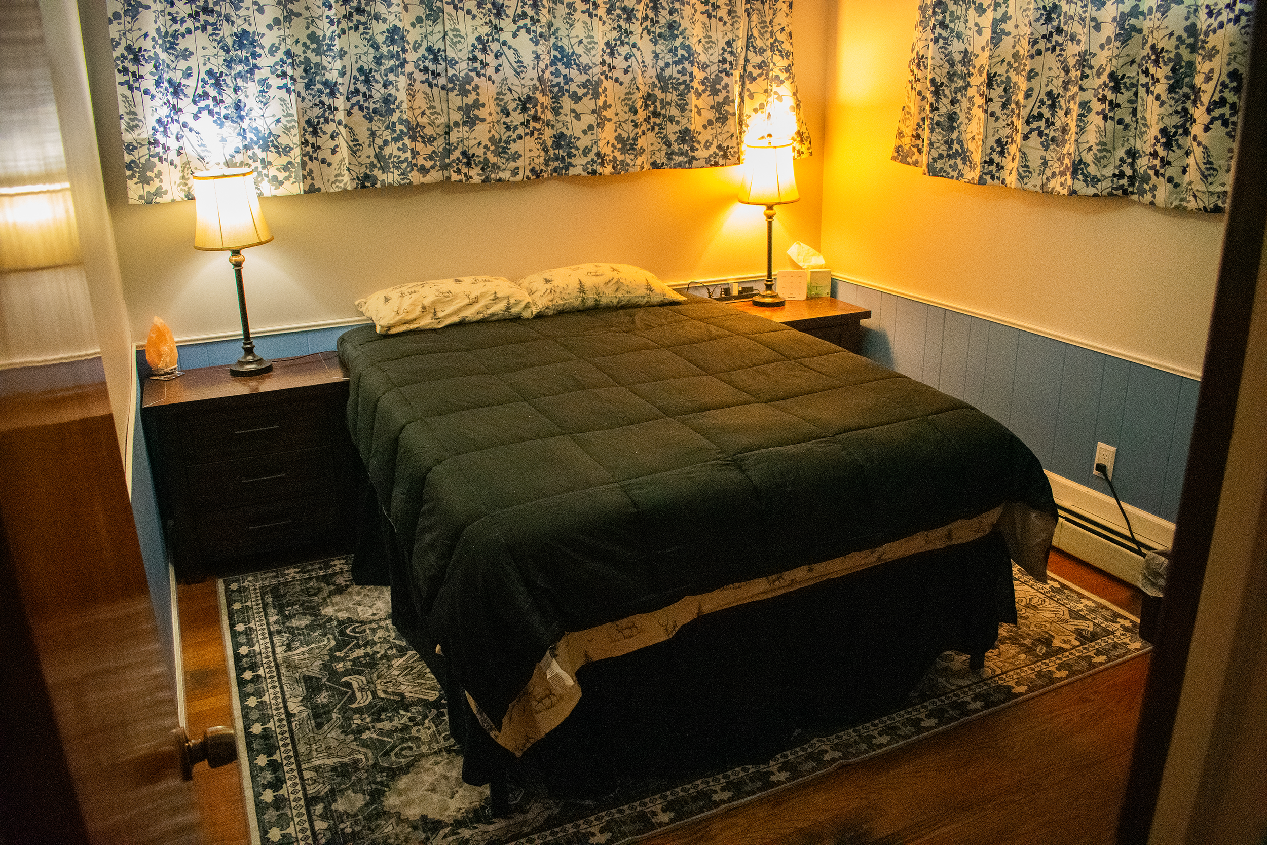 A bedroom with a wooden floor, a bed with dark blankets and a single pillow, two nightstands with lamps, a window with patterned curtains, and a ceiling fan with a colorful stained glass light fixture.