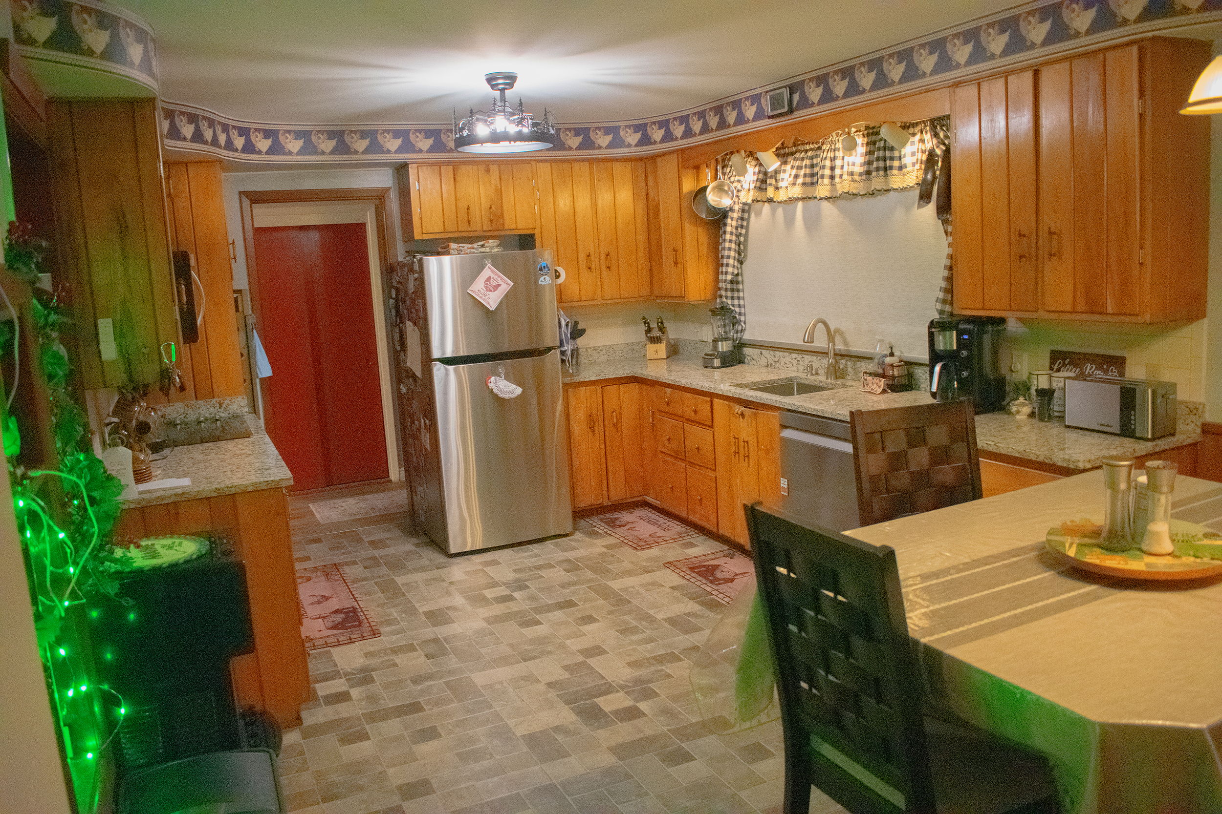 A cozy kitchen with wooden cabinets, granite countertops, and checkered curtains around the window. Stainless steel appliances include a refrigerator, dishwasher, and coffee machines on the counter. There are pots hanging above the sink, and a table with chairs in the foreground. The room has a tiled floor and decorative borders with rooster motifs.