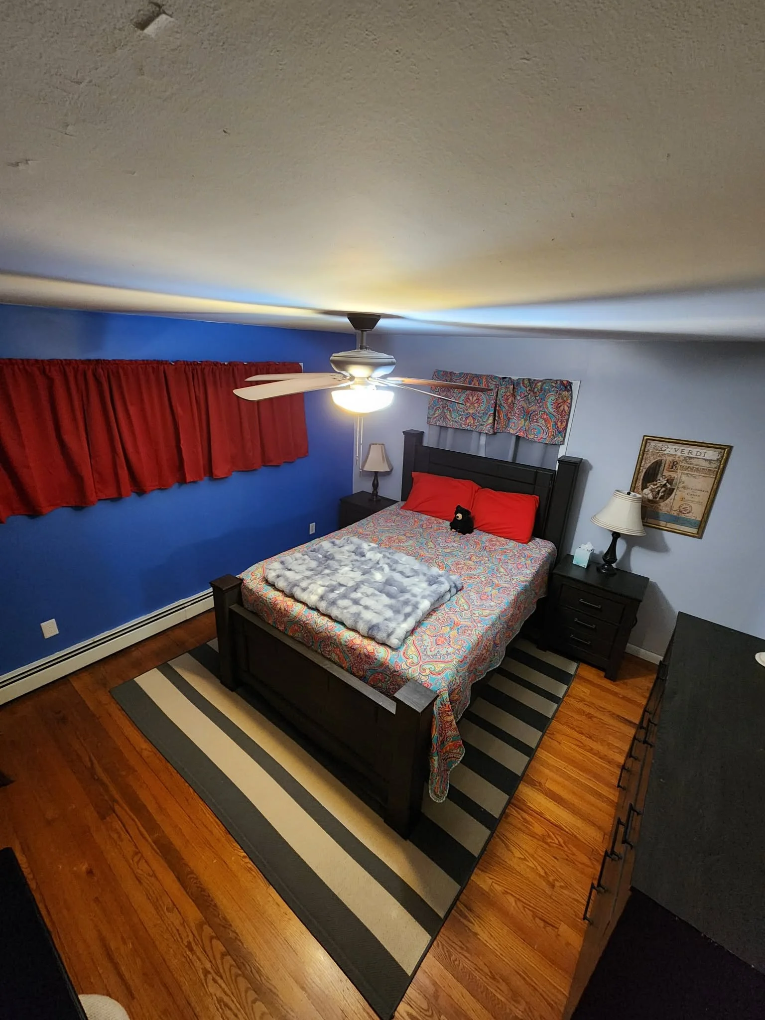 A bedroom with a wooden floor, dark furniture, a blue and red patterned bedspread, red pillows, and a small black stuffed animal. There are two nightstands with lamps, a framed picture on the wall, and a window with red curtains and a colorful valance. A ceiling fan lights the room.