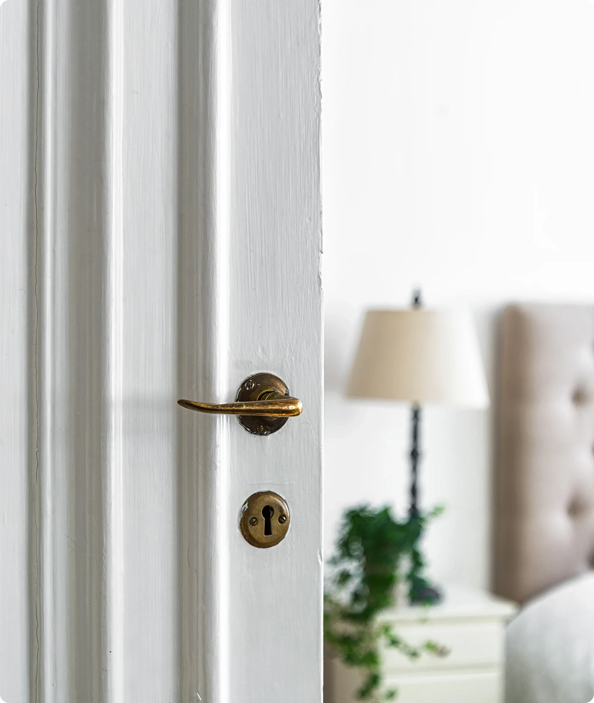 Close-up of a slightly open white door with a brass handle and keyhole, revealing a bedroom with a single lamp, a white nightstand, a framed picture on the wall, and a potted plant.