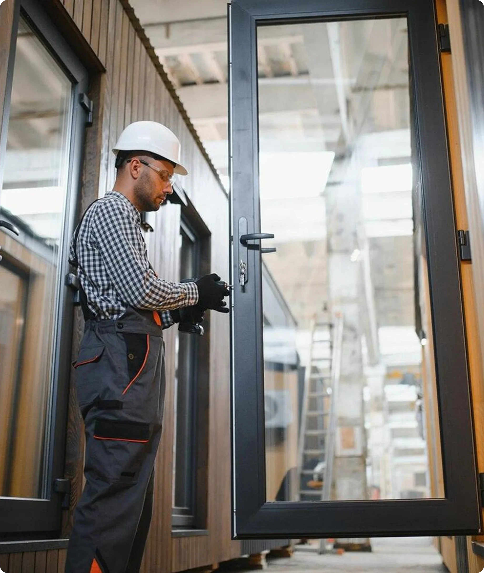 A man wearing a white safety helmet, gloves, and work clothes, holding a cordless drill, standing next to a glass door with a black frame at a construction site. The background shows other doors, building materials, and a ladder.
