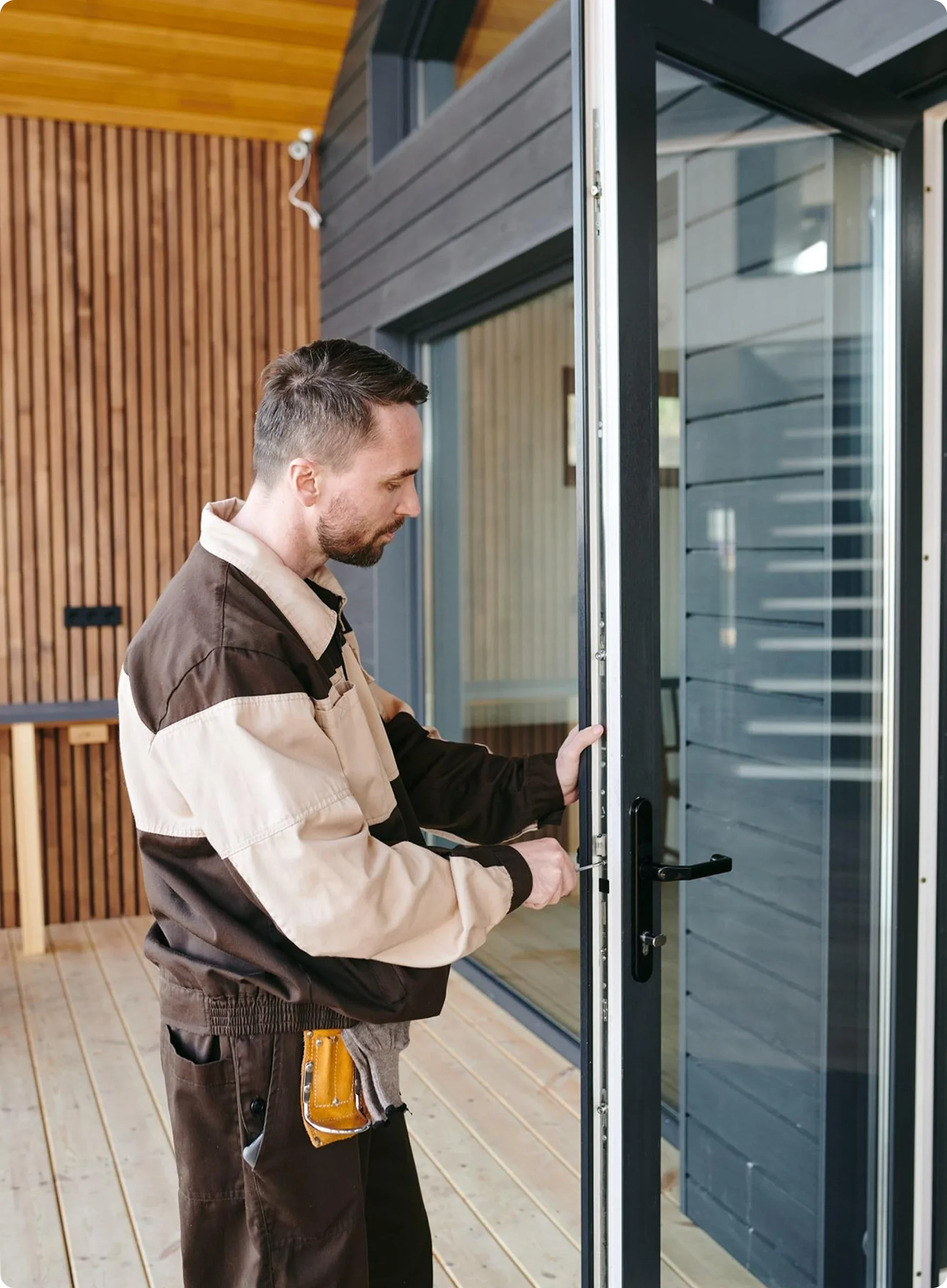 A man is installing or repairing a glass door, wearing a beige and black uniform with a pliers tool in his pocket, on a wooden deck of a modern house with blue-gray siding.