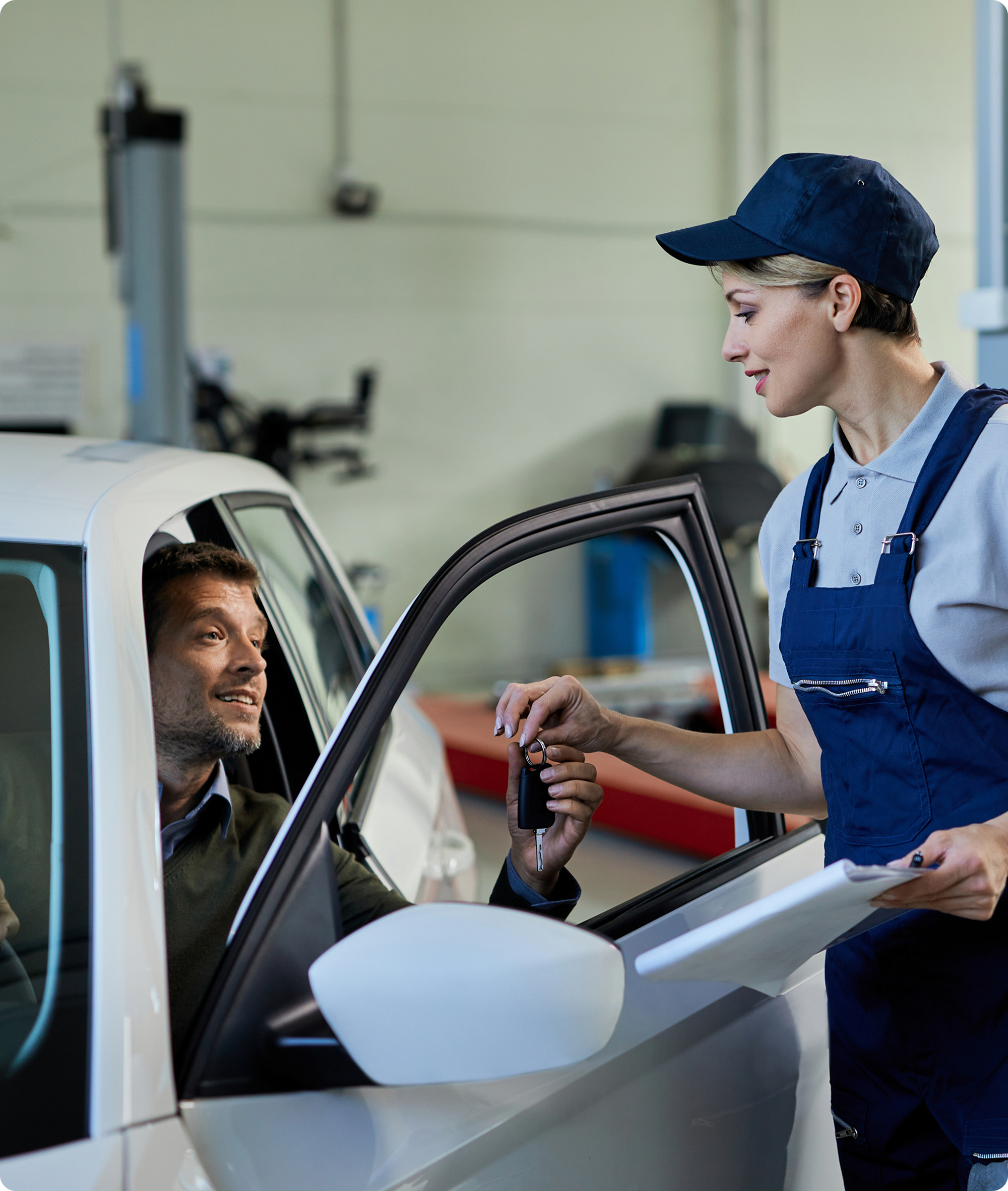 A woman in a blue uniform and cap hands car keys to a man sitting in a white car while holding a clipboard, inside an auto repair shop.