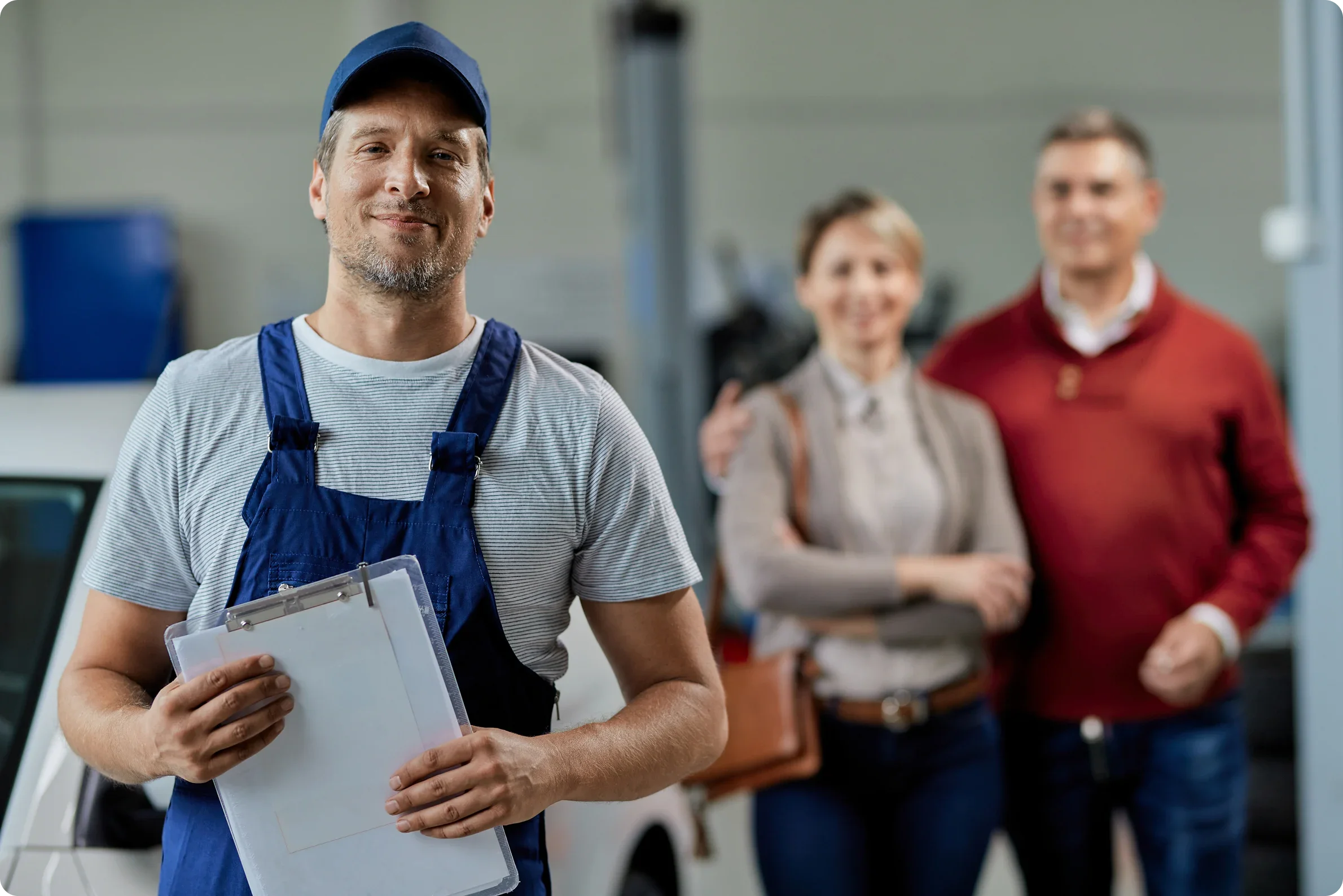 A man in an apron holding a clipboard, smiling at the camera, with two people in the background, a woman and a man, standing close together and smiling.