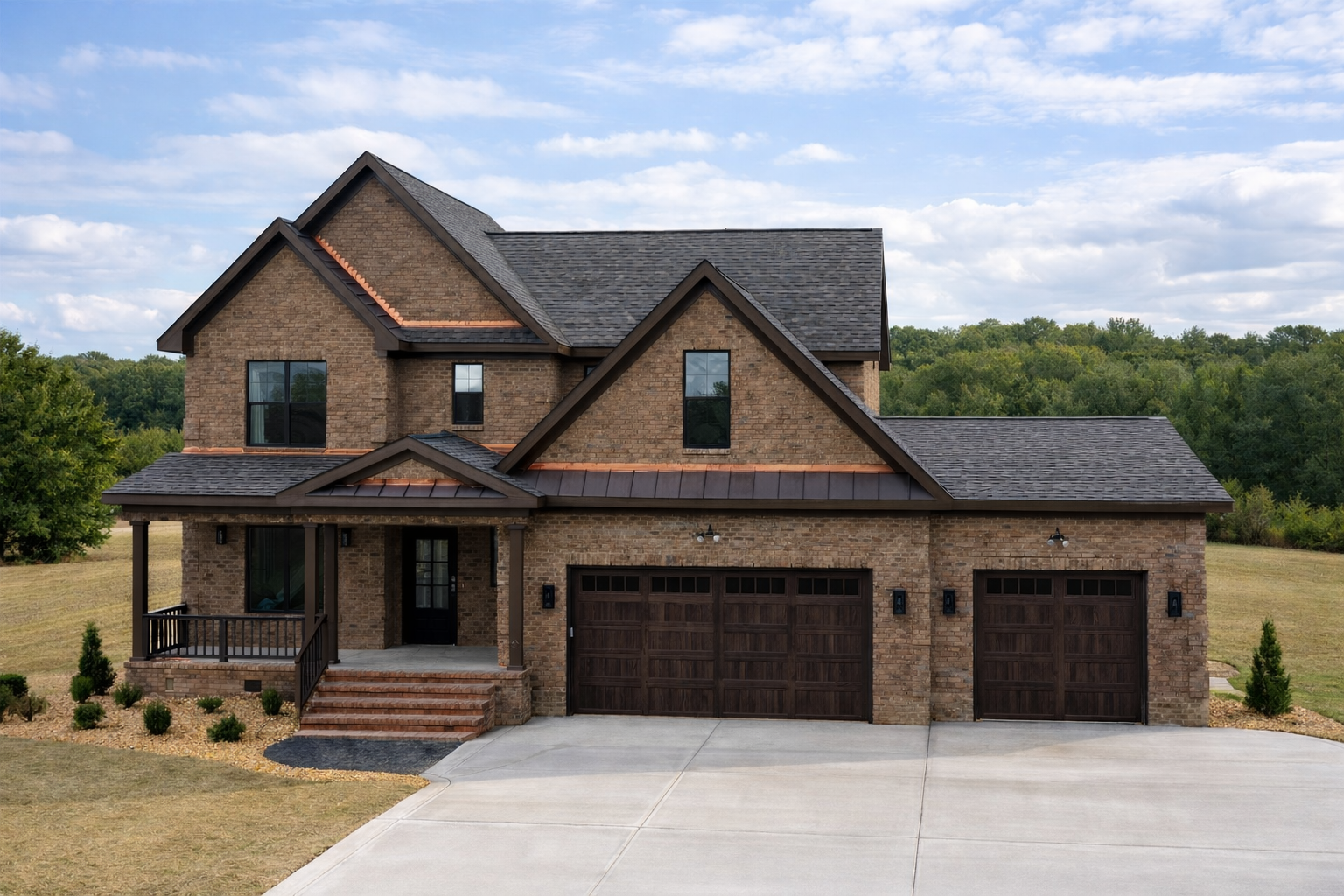A two-story brick house with a dark shingle roof, two garages with wooden doors, a small front porch with steps, and a landscaped yard with shrubs and small trees, set against a background of green trees and a partly cloudy sky.