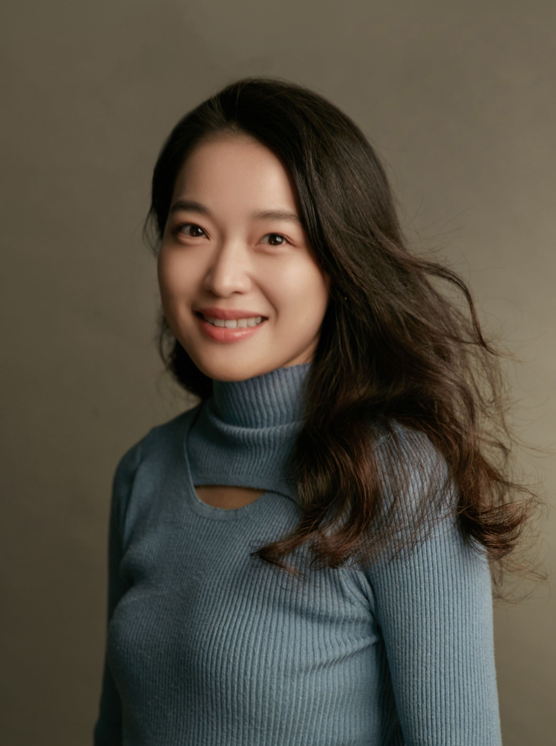 一名留著長長波浪狀黑髮的女子，身穿淺藍色羅紋高領毛衣，面帶微笑，背景為素雅的米色。