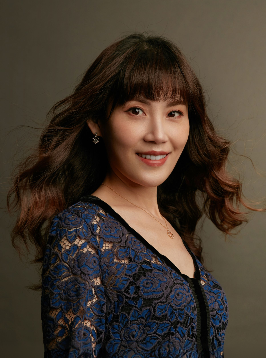 一名女子的肖像：她留著一頭長長的棕色捲髮，身穿藍黑相間的蕾絲上衣，配戴耳環與項鍊，在灰色背景前展露笑容。