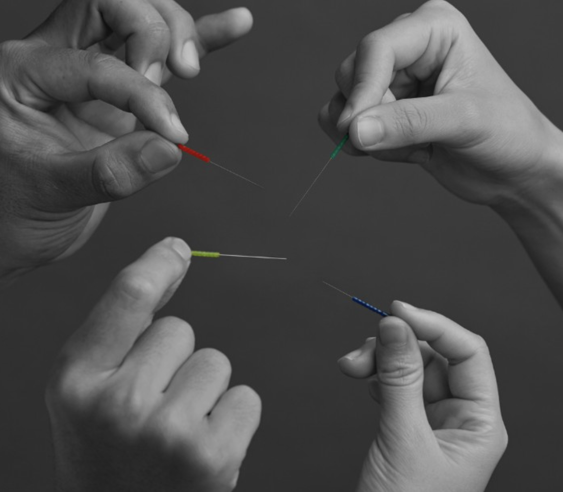 Four hands holding acupuncture needles with colored tips, arranged in a circle against a dark background.