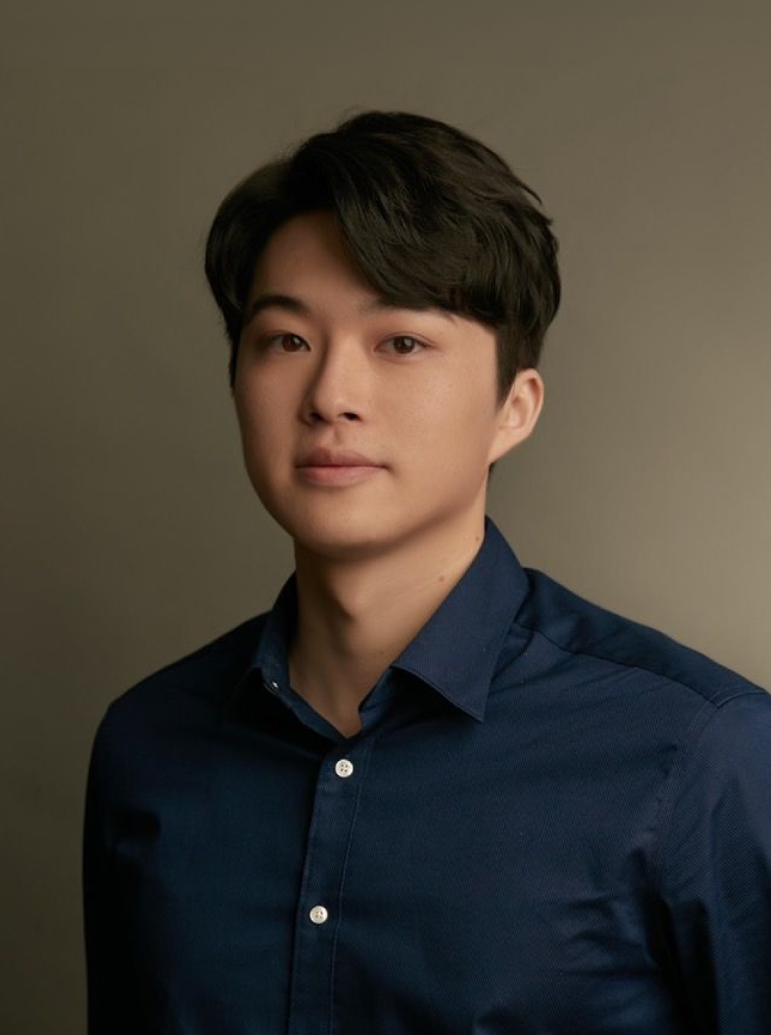 一名黑髮青年身穿海軍藍鈕扣襯衫，站在素色背景前的肖像。