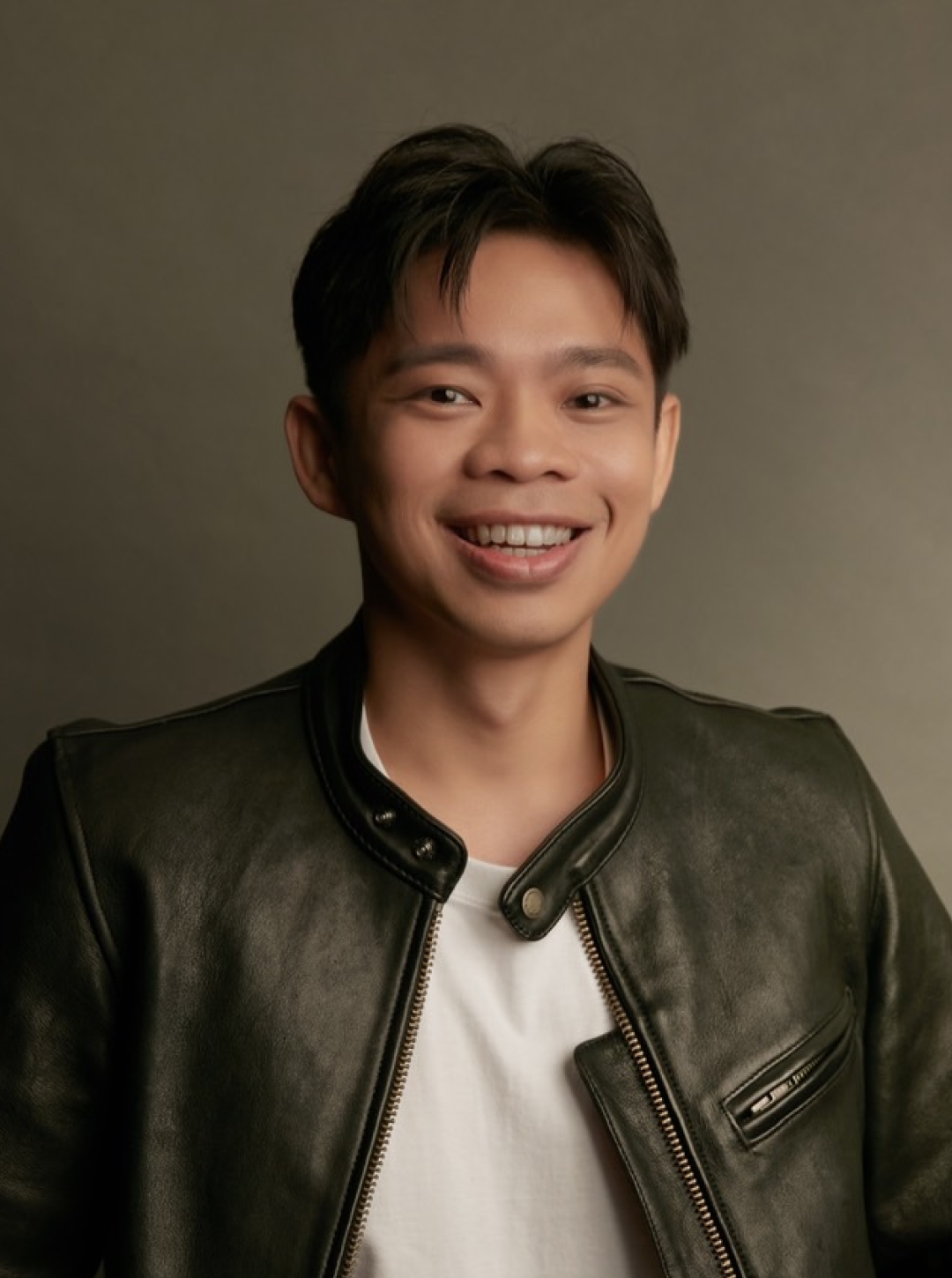 一名年輕的亞洲男子面帶微笑，身穿黑色皮夾克，內搭白色T恤，站在素色背景前。