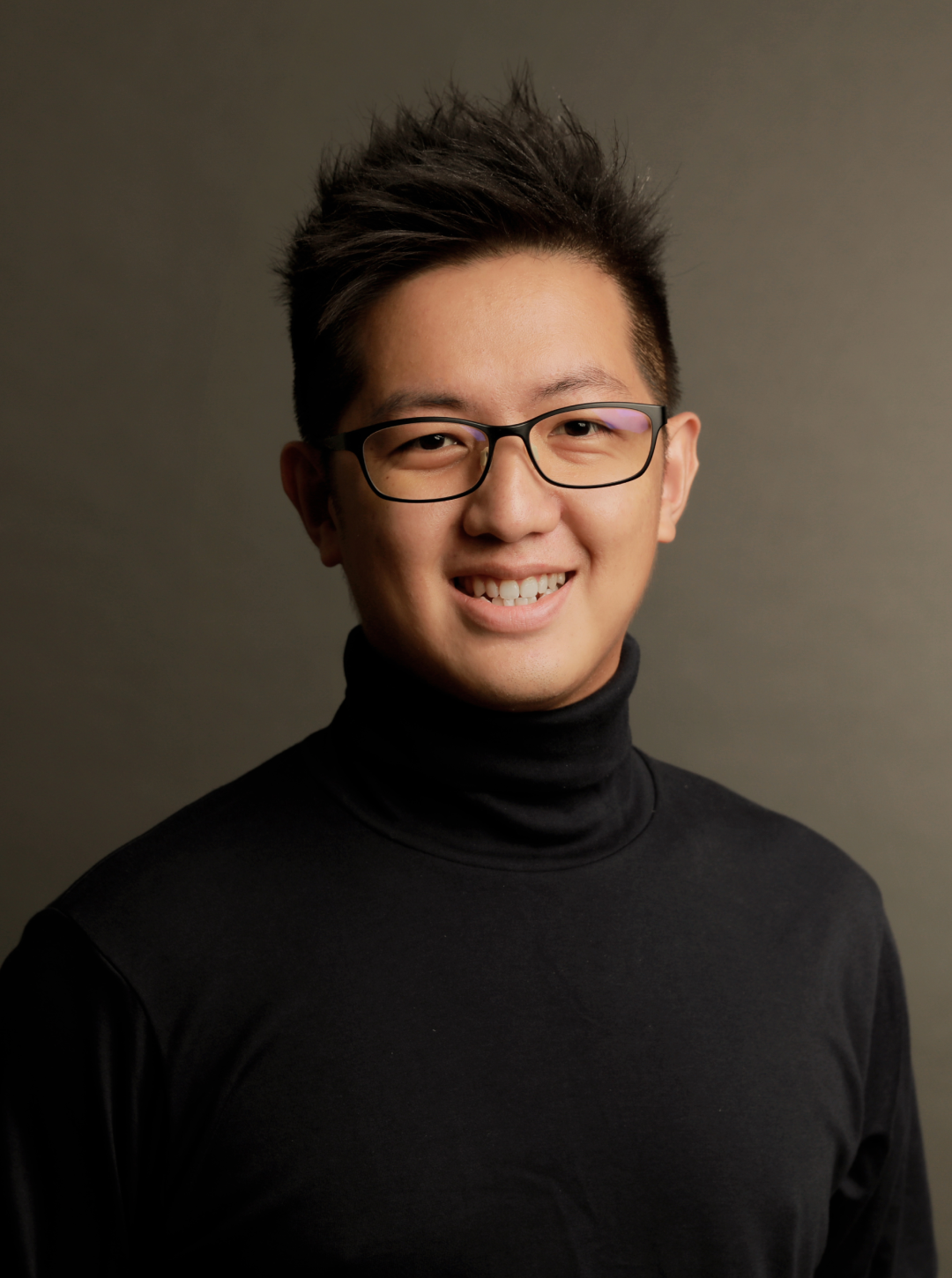 一名戴著眼鏡、身穿黑色高領衫的年輕男子，在素色背景前微笑的半身照。