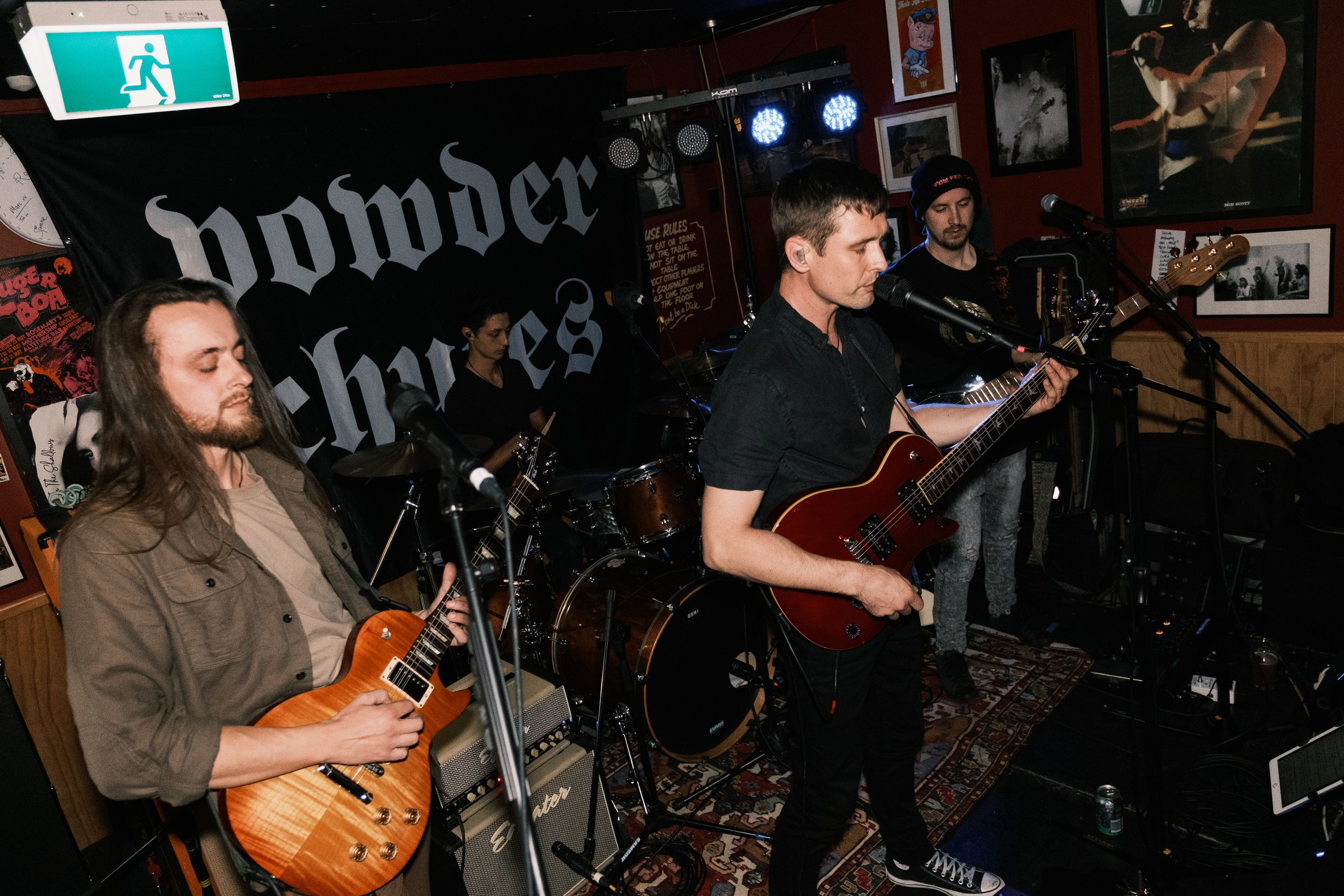 A band performing live in a dimly lit venue with posters and framed photos on the walls, and a green emergency exit sign visible at the top left.
