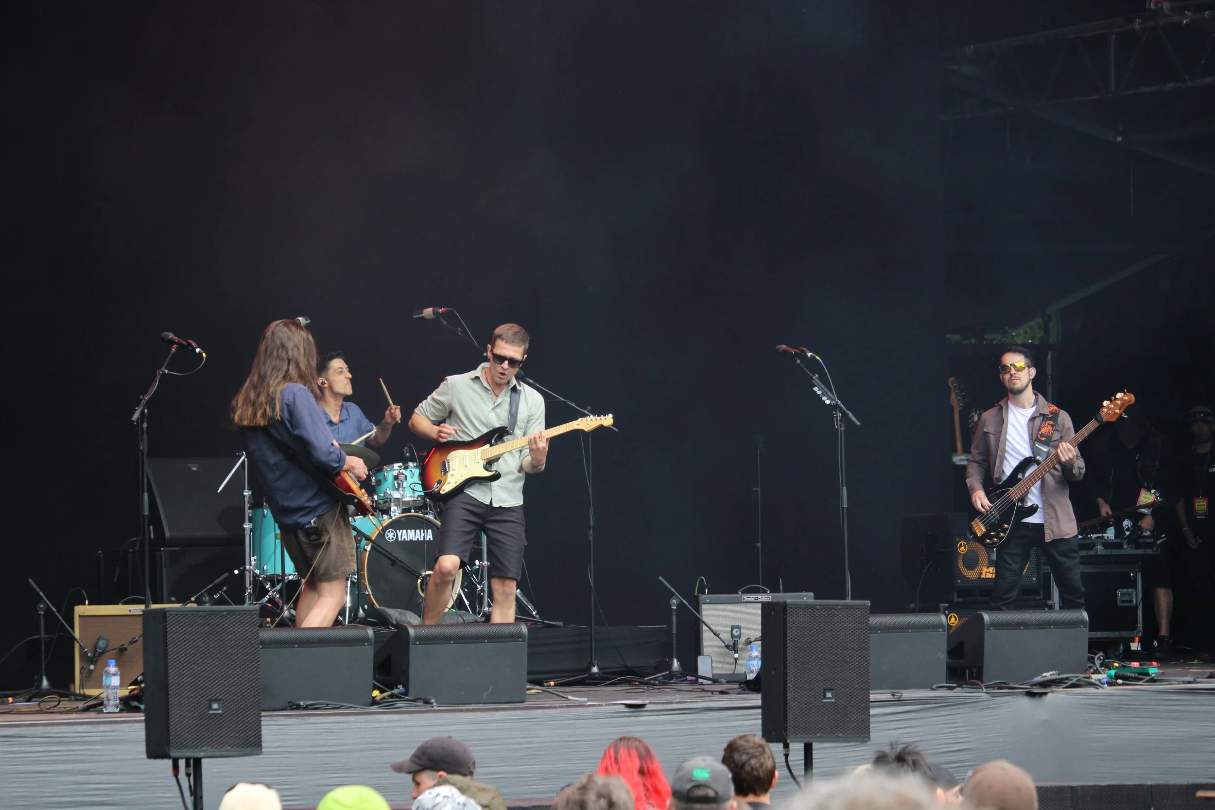 A band performs on stage at an outdoor concert, with members playing guitars, drums, and bass, and people in the audience watching.