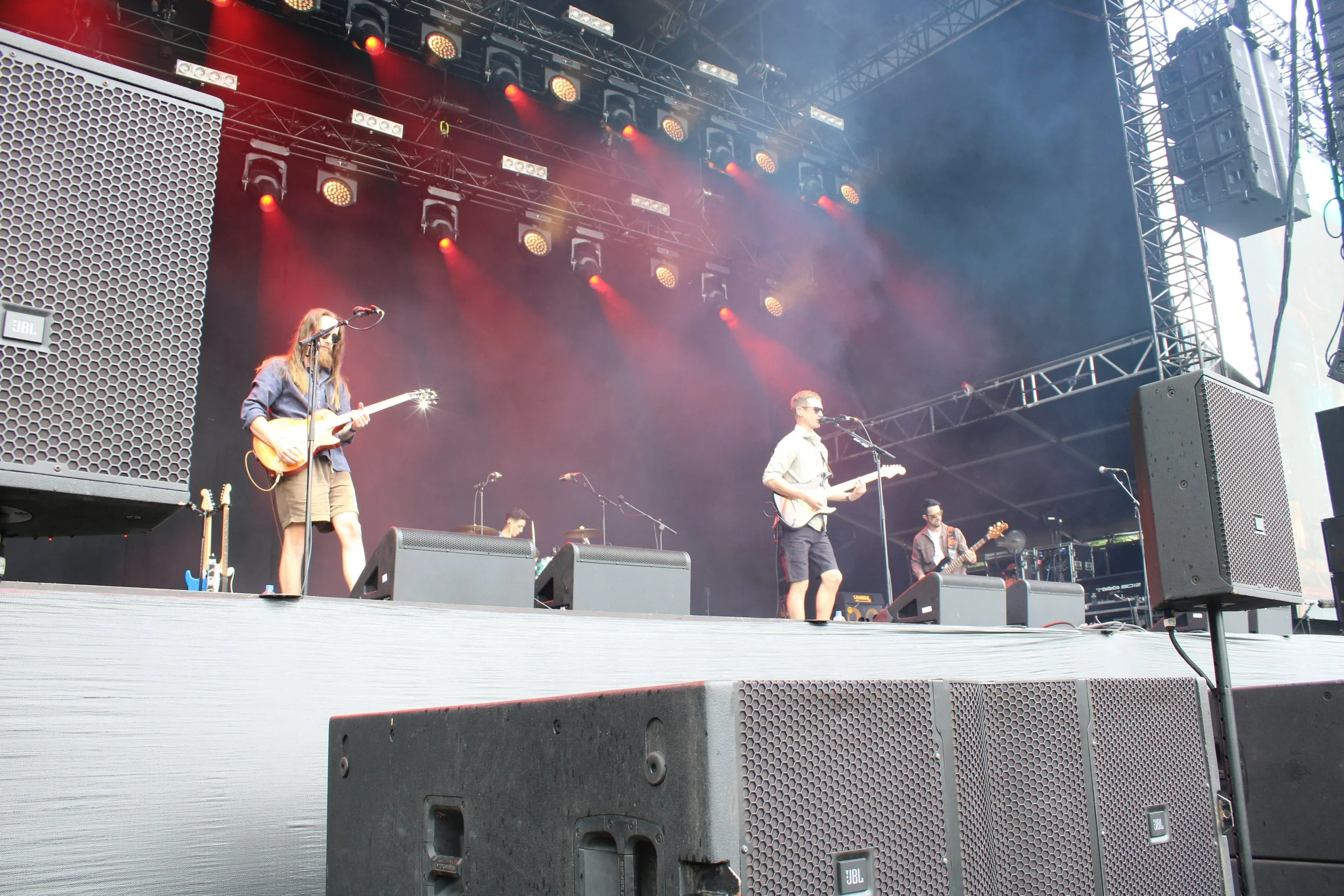 A band performing on an outdoor stage with musical instruments, microphones, and large speakers, under stage lighting and equipment.