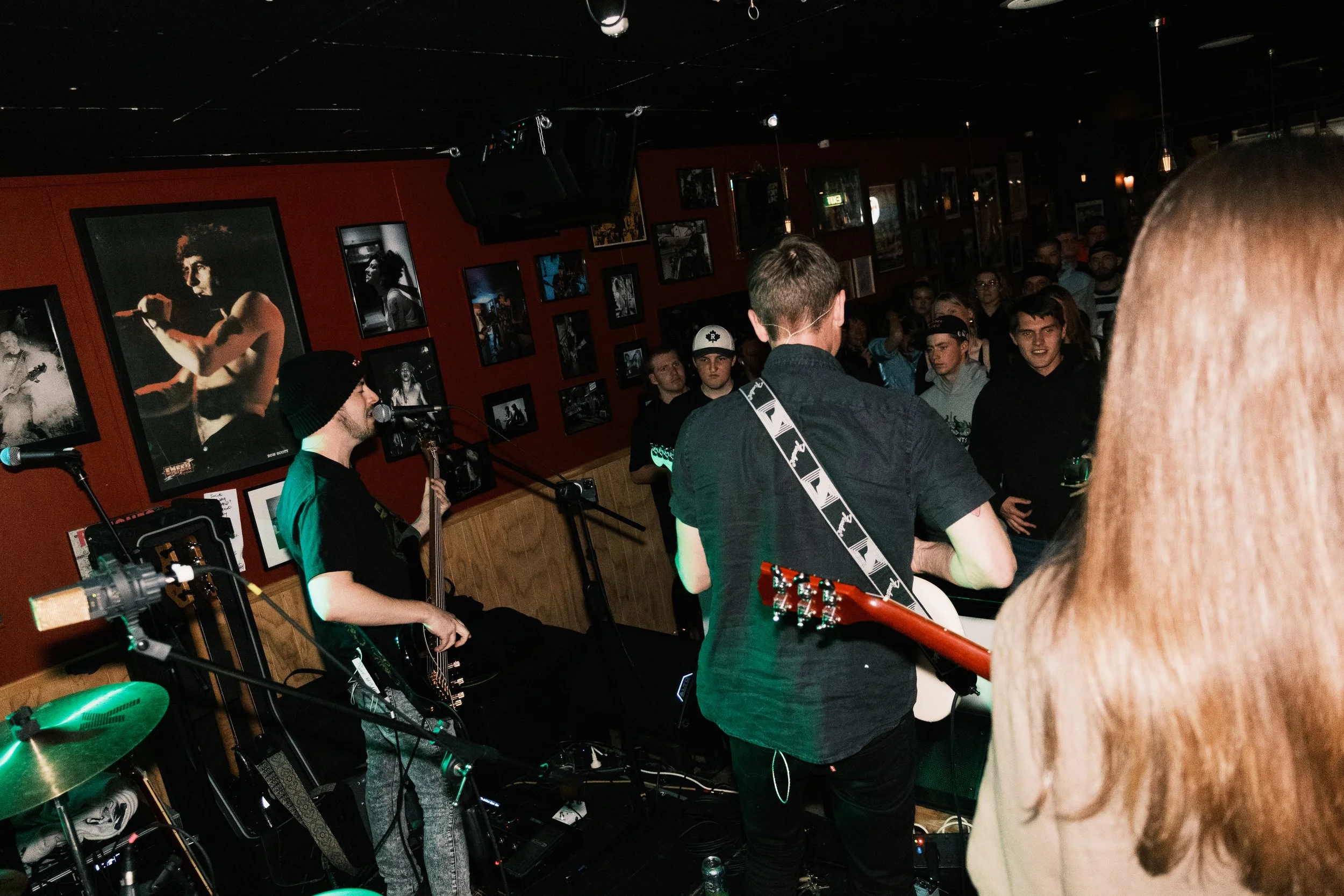 A live music performance with a singer and a guitarist in a dimly lit venue, surrounded by an audience. The venue has black walls decorated with framed photos, and there are spotlights on the performers.