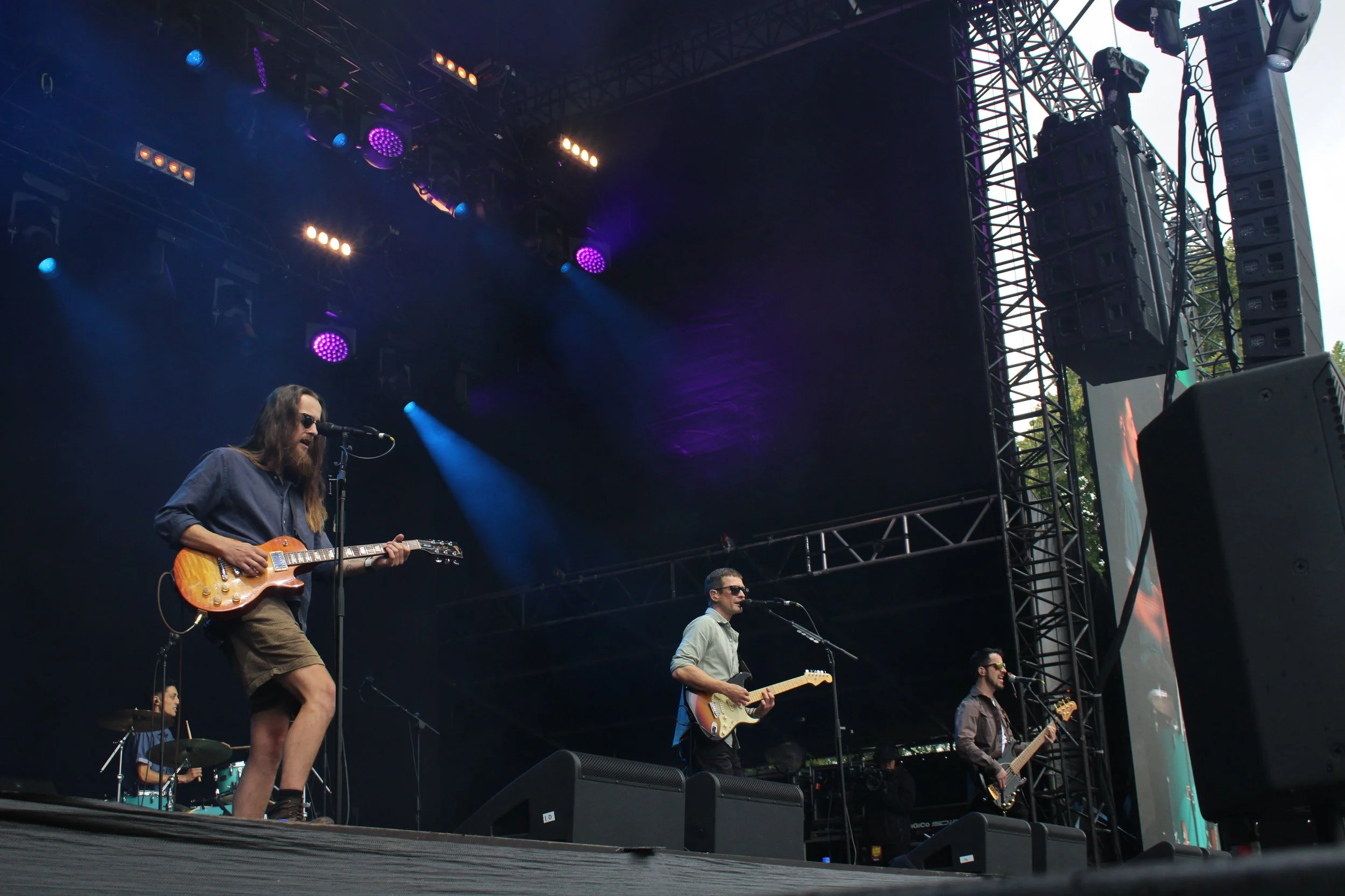 Musicians performing live on an outdoor stage during a concert, with colorful stage lights, large audio speakers, and a screen displaying visuals.