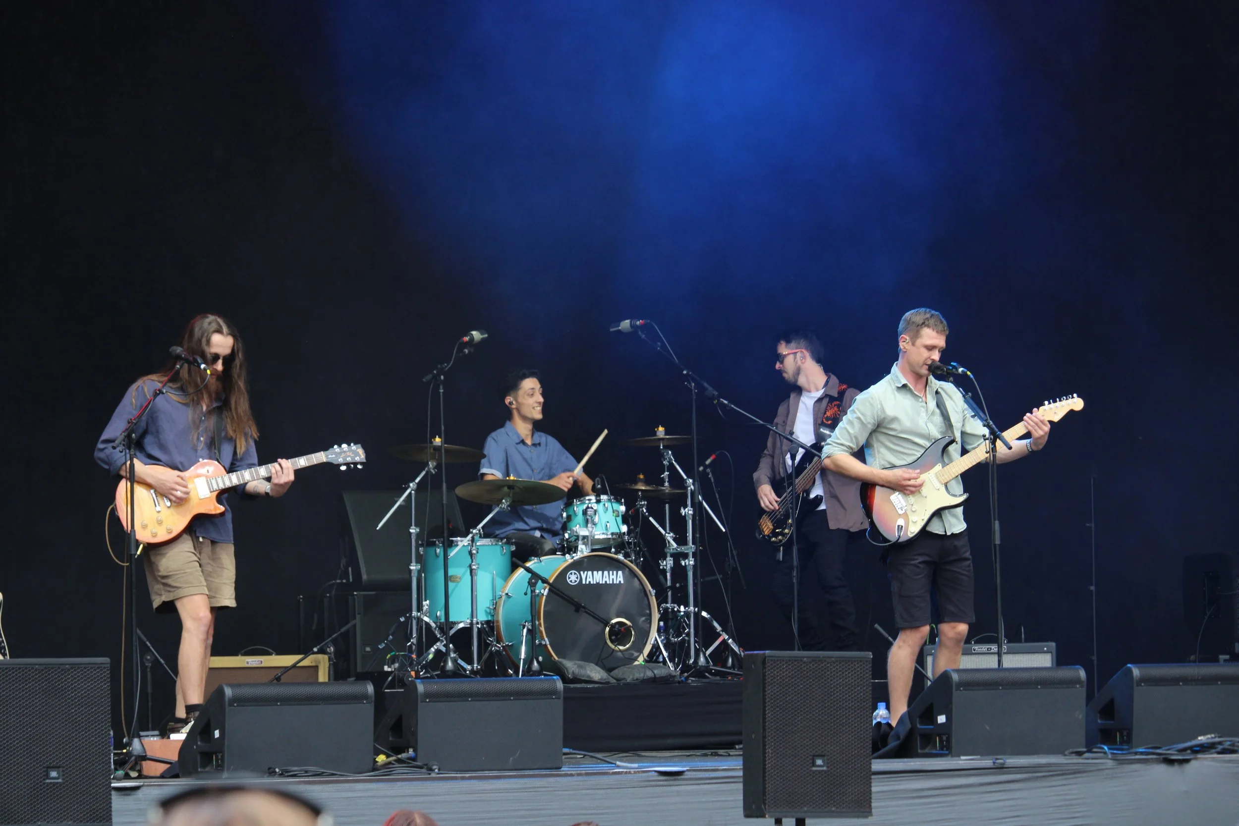 A band performing on stage with four members playing instruments: an electric guitar, drums, bass, and another electric guitar, against a dark background with blue lighting.