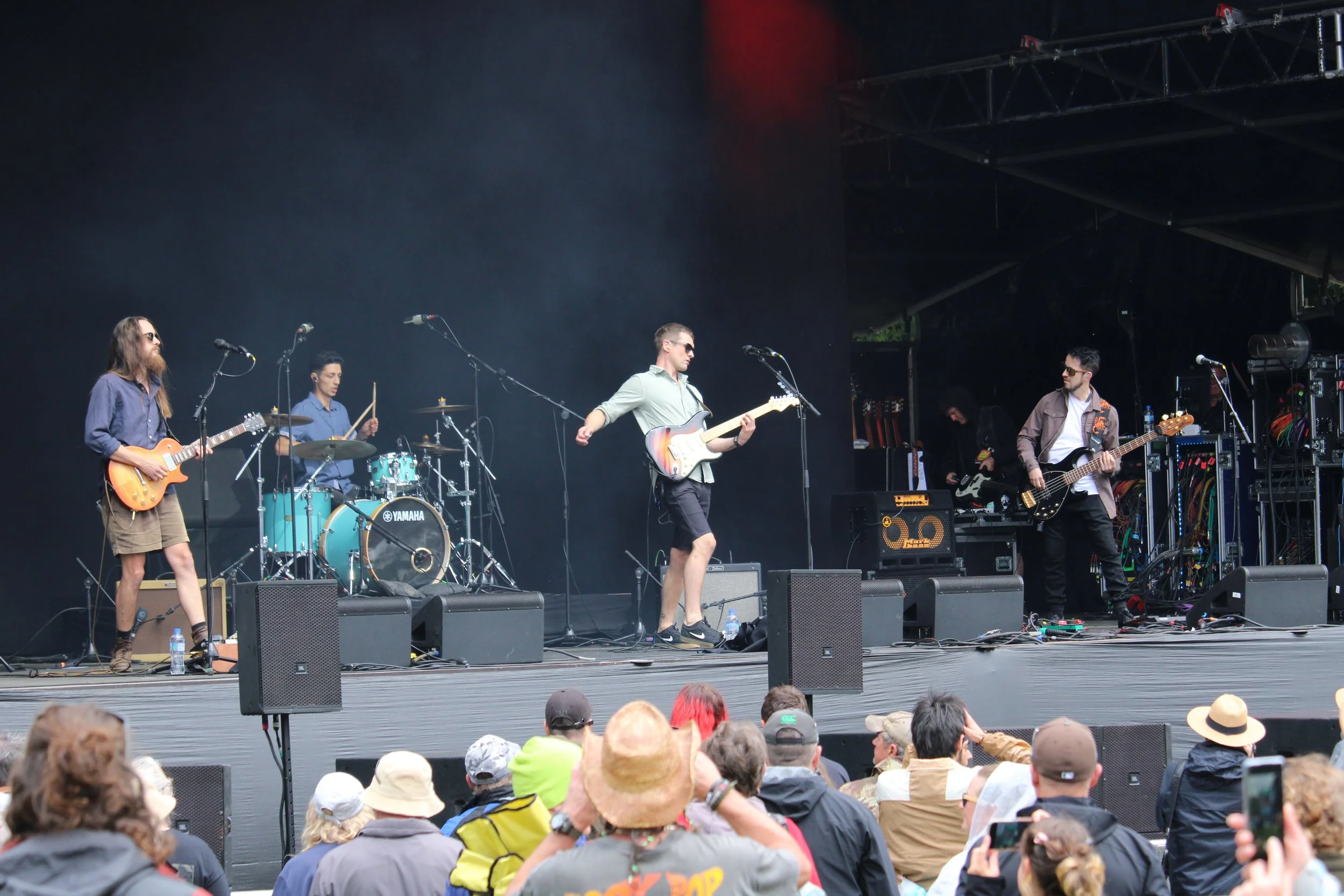 A band performing on an outdoor stage with musicians playing guitars, drums, and bass guitar, while an audience watches.