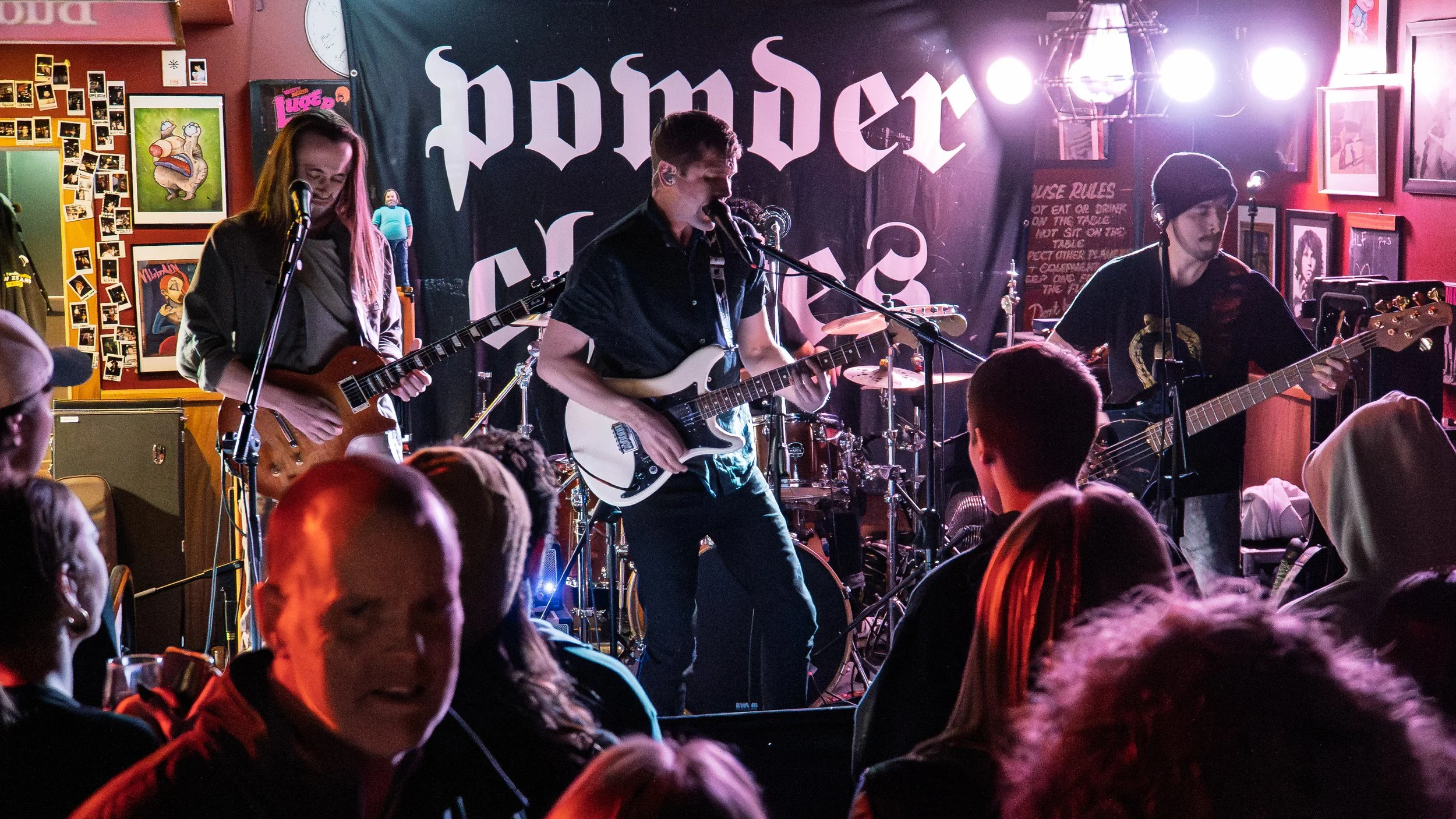 A band performs live music on stage in a bar or club, with a backdrop reading 'Homer'. The audience is visible in the foreground, and the scene is lit with colorful stage lighting.