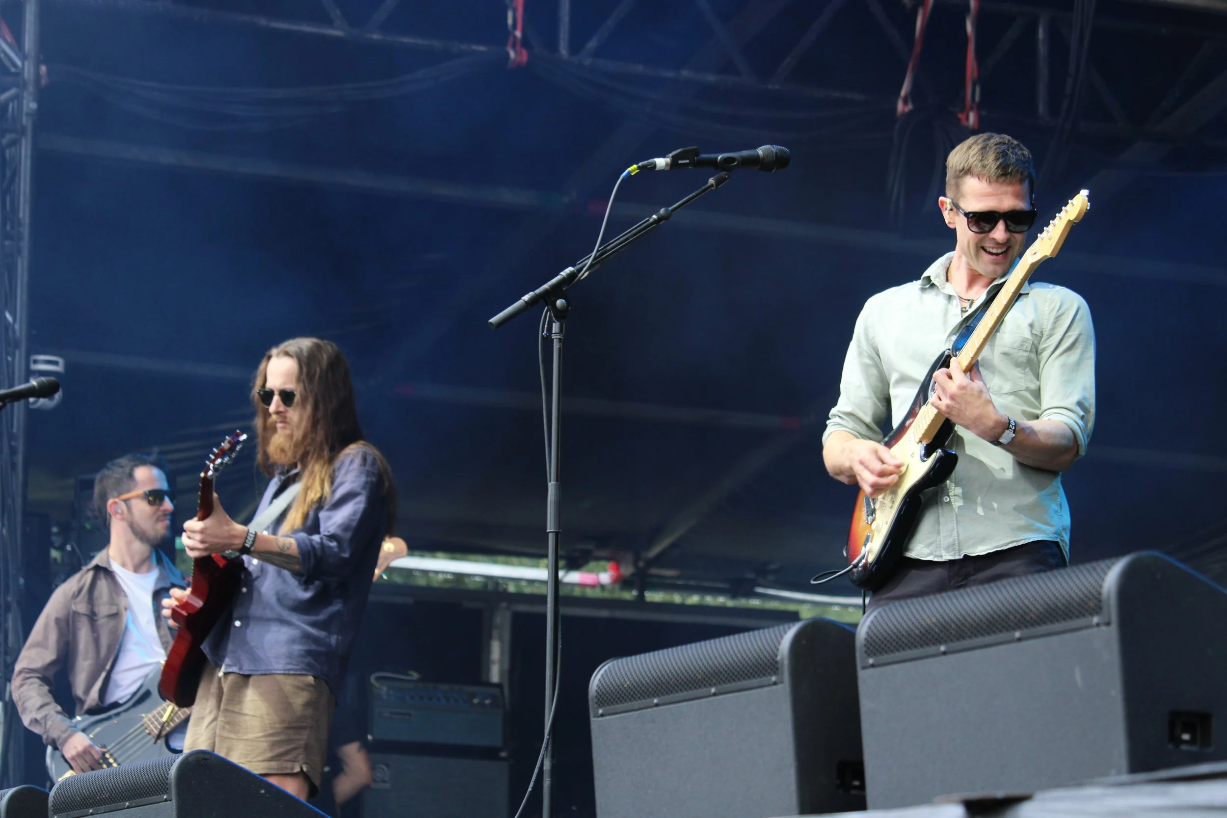 Musicians performing on an outdoor stage, with one man playing an electric guitar and wearing sunglasses, while a woman playing a guitar stands nearby. There are other musicians and audio equipment visible.