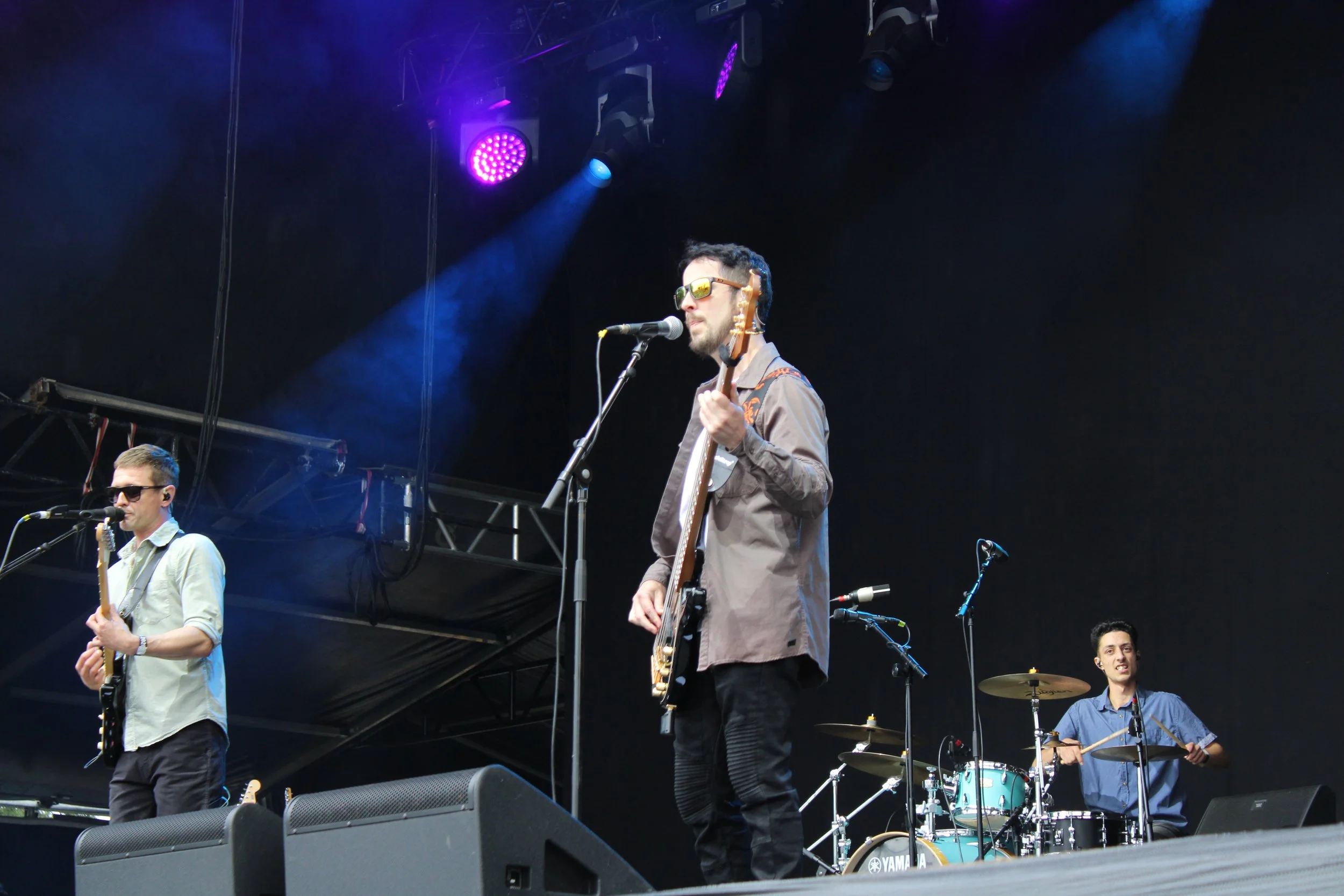 Four band members perform on stage during a concert: a man with sunglasses singing into a microphone while holding a guitar, a man in a light shirt playing a guitar, another man with sunglasses standing behind a microphone, and a drummer in a blue shirt playing drums. There are purple stage lights overhead.