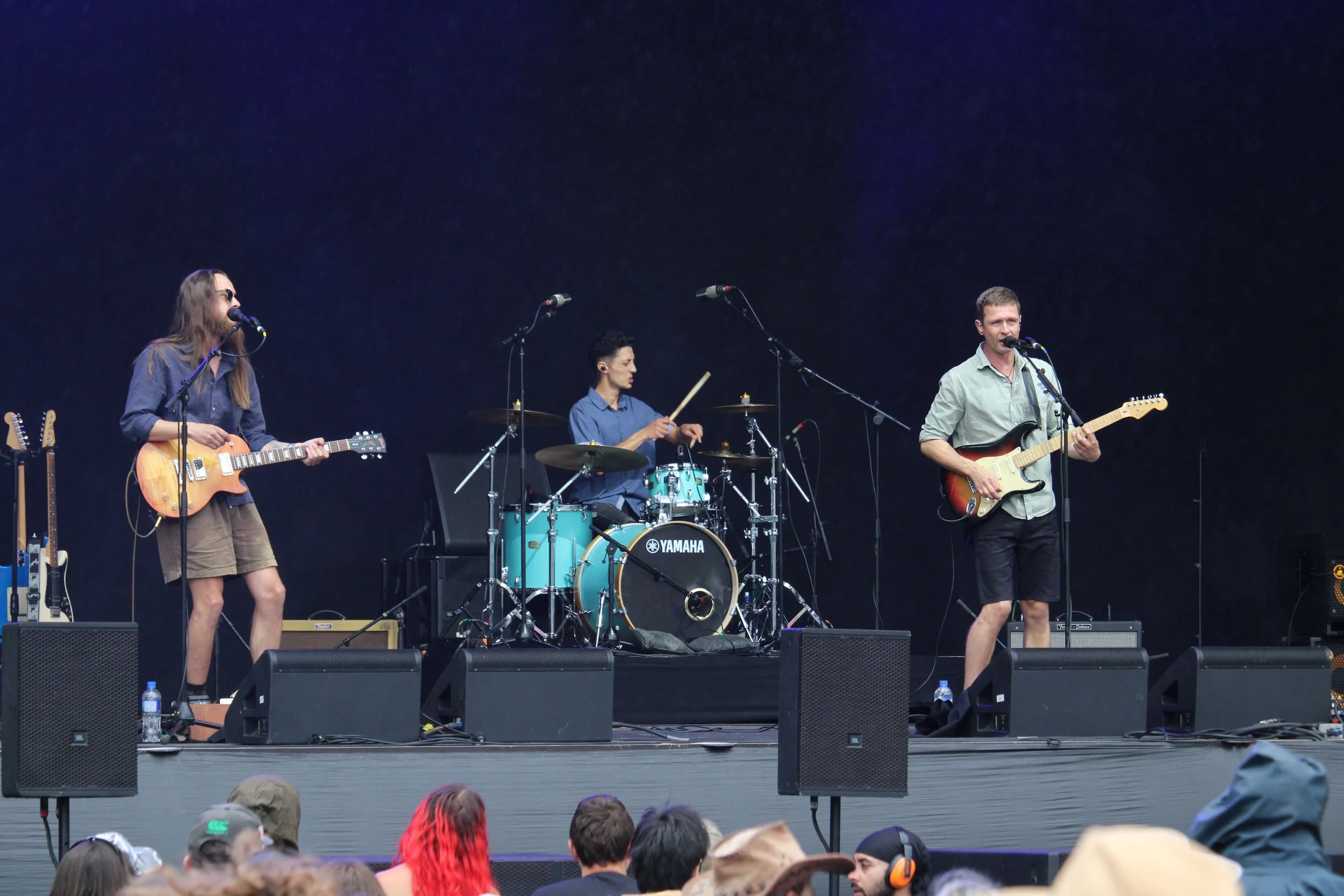 A band performing on stage at an outdoor concert, with a woman playing guitar and singing, a woman on drums, and a man playing electric guitar and singing.