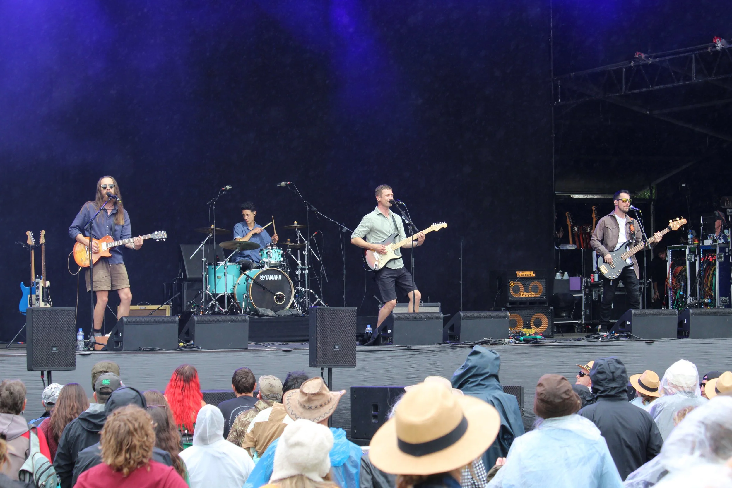 A band performing live on an outdoor stage with audience members watching, some wearing hats and jackets.