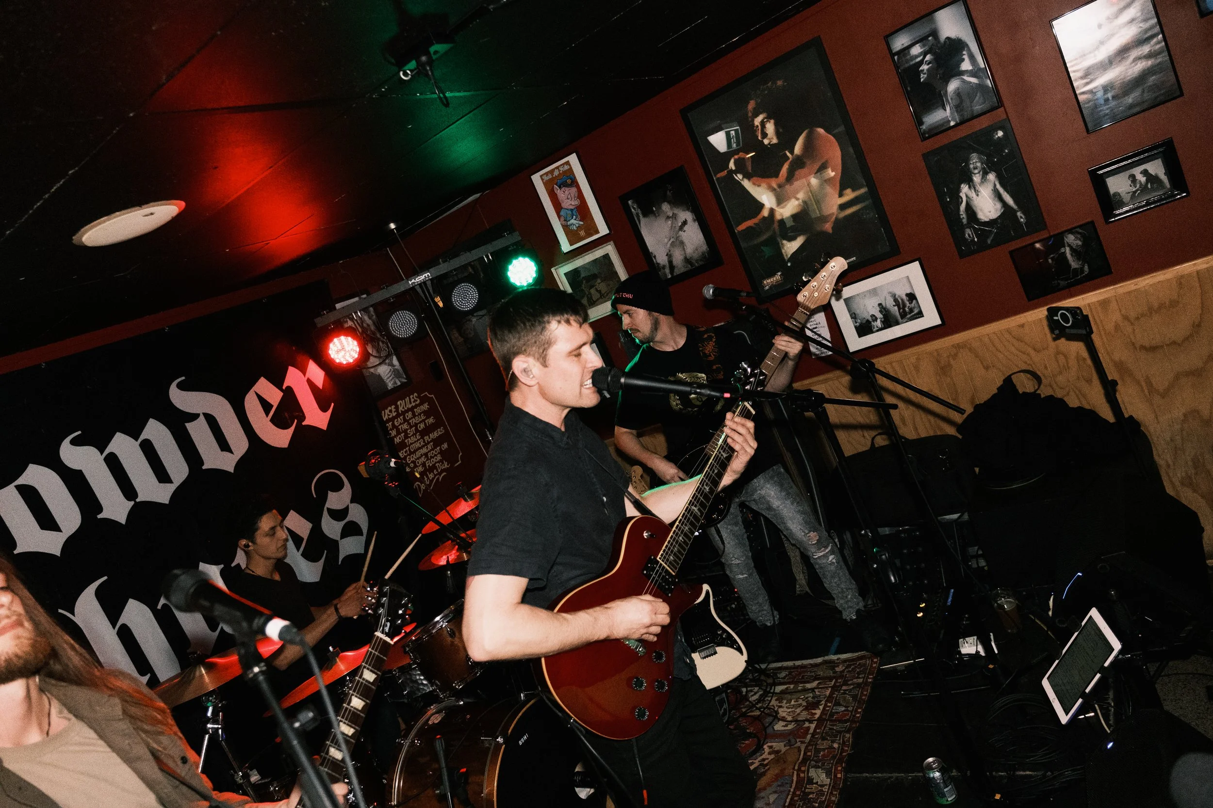 A live band performs on a small stage in a dimly lit venue with photos on the wall and red and green stage lights. The band includes a vocalist or guitarist singing into a microphone and a guitarist playing. An audience is partially visible.
