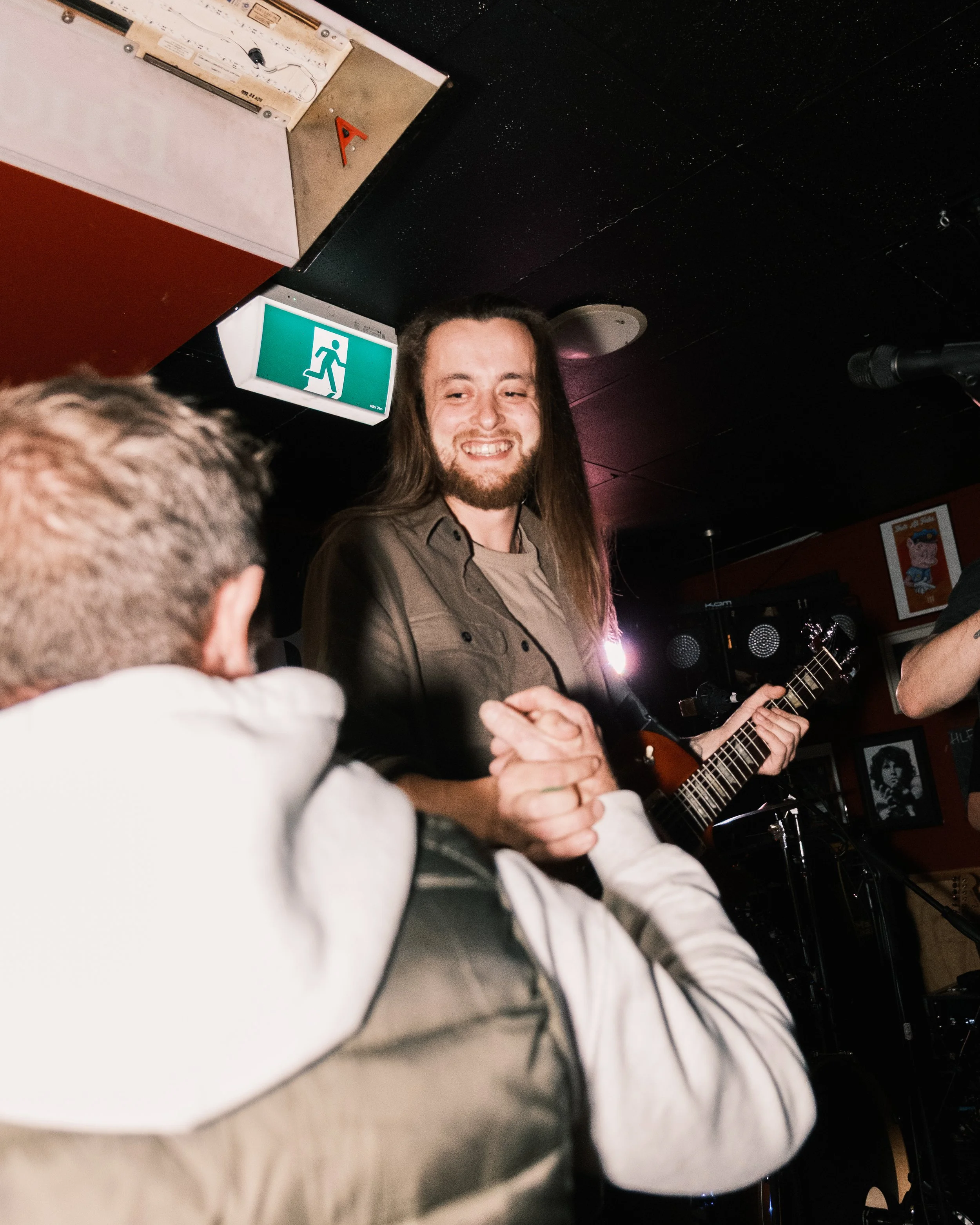 A man with long hair and a beard playing an acoustic guitar, smiling, in a dimly lit indoor setting with posters on the wall and a microphone nearby.