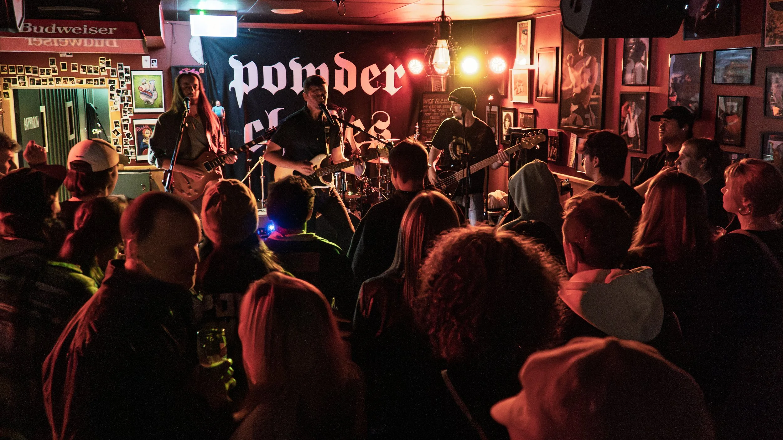 A band performing live on stage in a dimly lit venue with an audience watching. The stage has musical instruments, and the background features a dark curtain with some text.