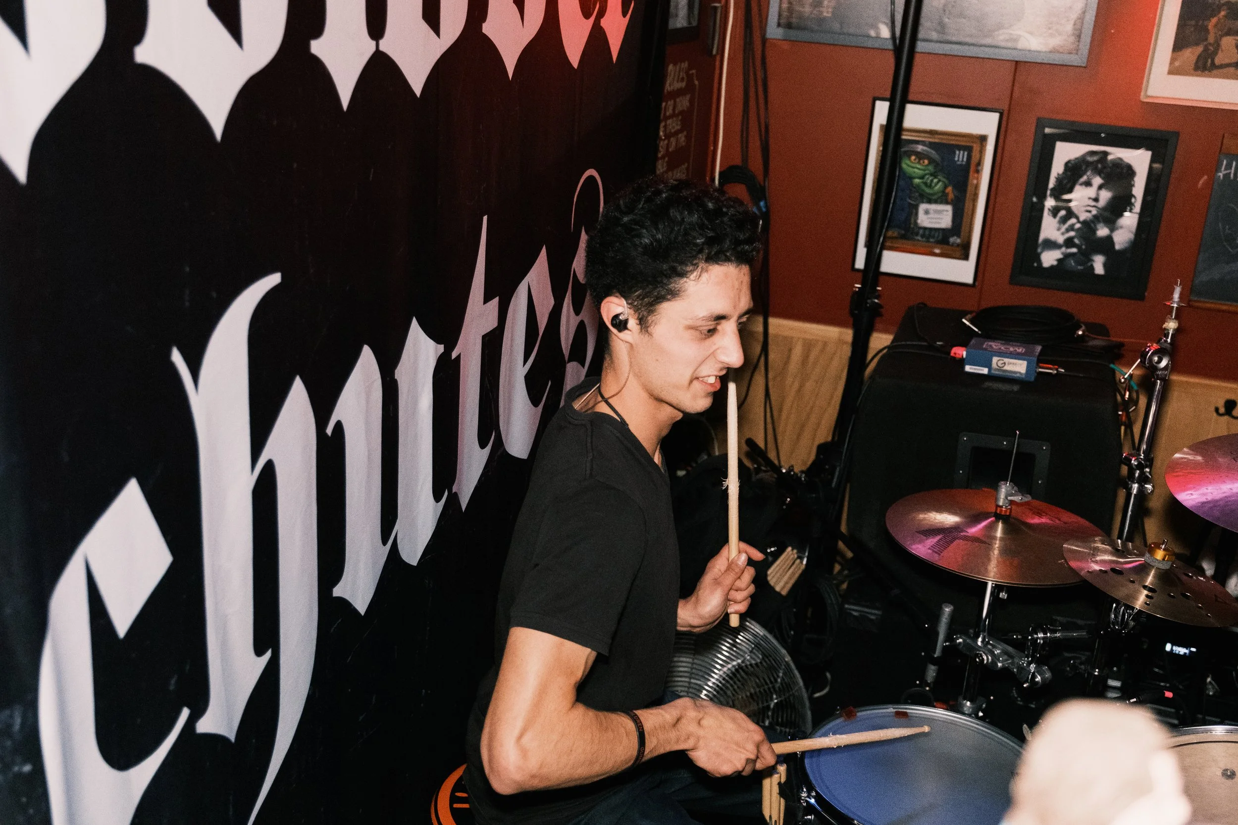 A young man playing drums in a music venue. He's holding drumsticks, and there are framed photos on the wall behind him, including a black and white photo of a woman and a colorful frog illustration.