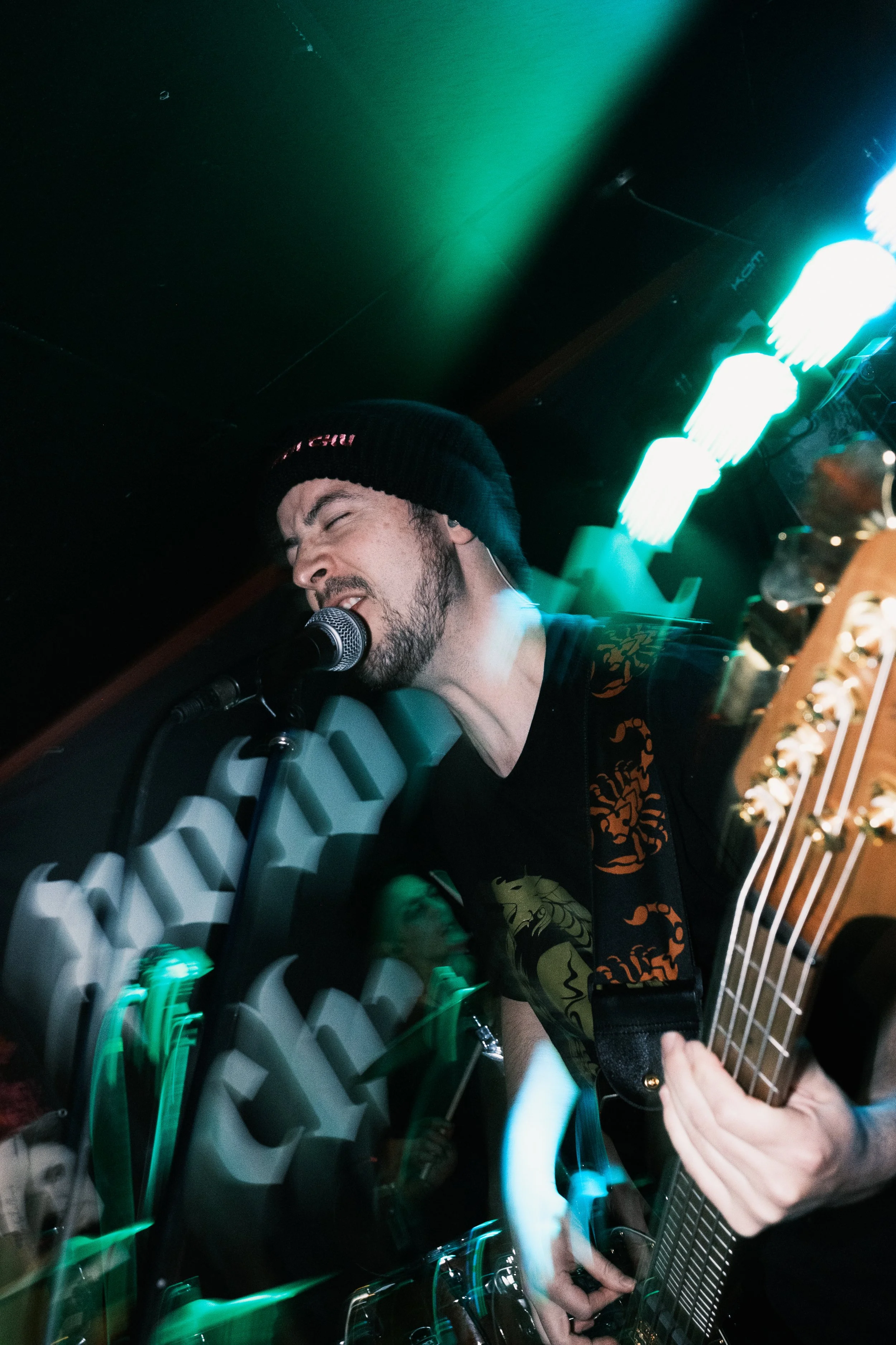 A person wearing a black beanie and a black shirt is singing into a microphone while playing an acoustic guitar. The background features illuminated green and white lights, creating a vibrant atmosphere.