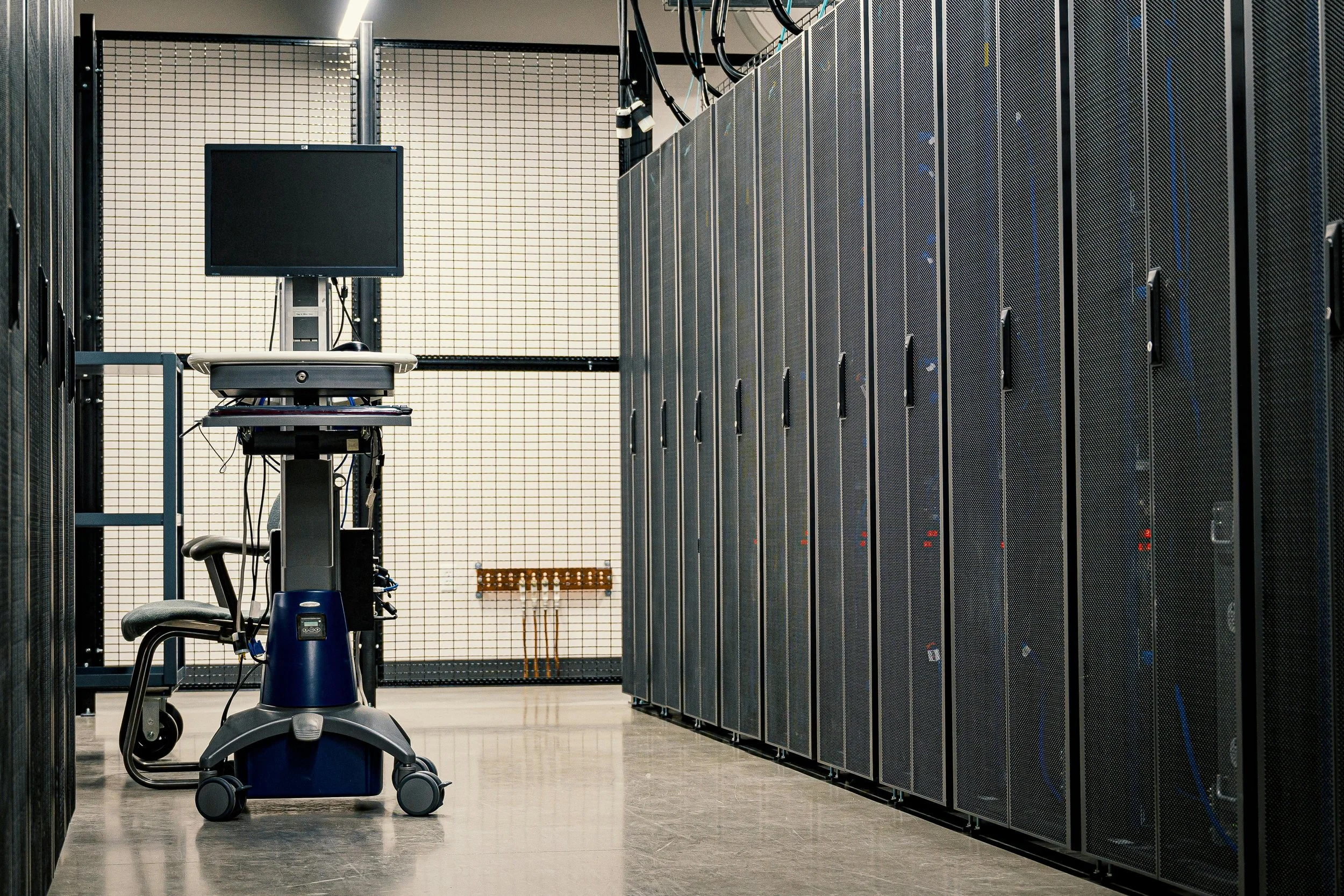 Data center room with black server racks on the right, a mobile computer station with monitor, keyboard, mouse, and a rolling chair on the left, and a perforated metal grid wall in the background.