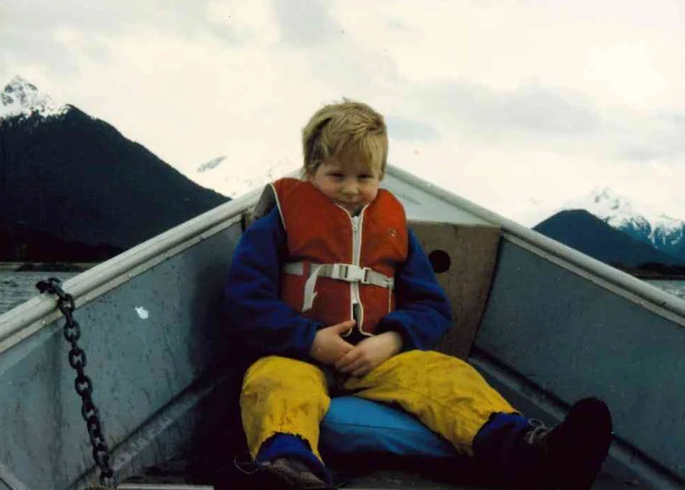 Young JKT wearing a red life jacket, blue jacket, yellow waterproof pants, and black boots sitting in a small boat with a snowy Alaskan mountain landscape in the background.