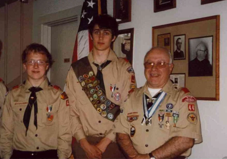 JKT dressed in an Eagle Scouts uniform, wearing badges and neckerchiefs, standing indoors in front of a framed photo collage and an American flag.