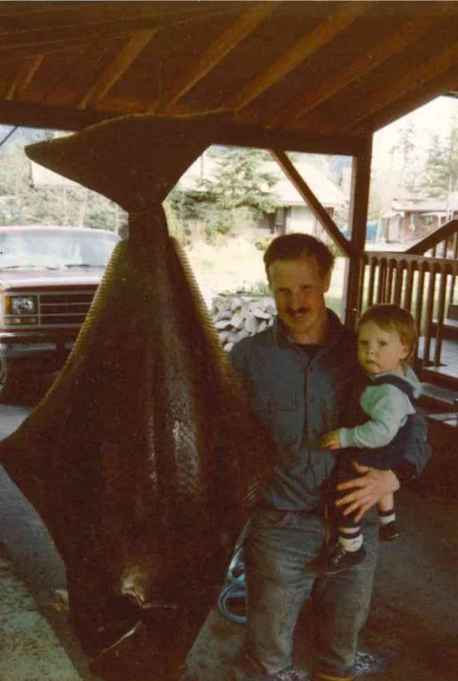 A man holding a young JKT standing next to a large fish hanging from a ceiling in a rustic outdoor setting.